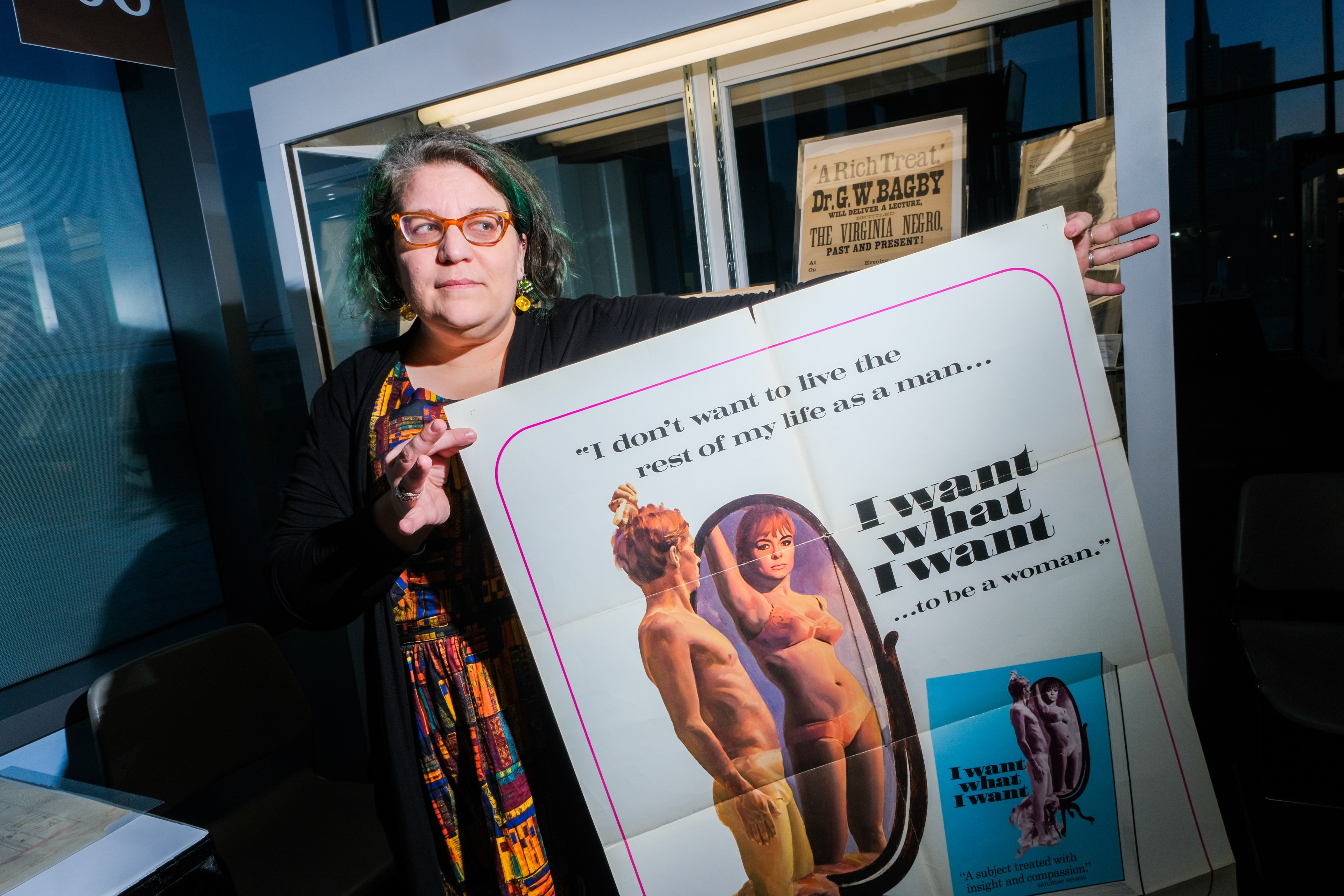 A person holds a large poster with text and an illustration of a nude figure transitioning genders. The poster has a feminist message.