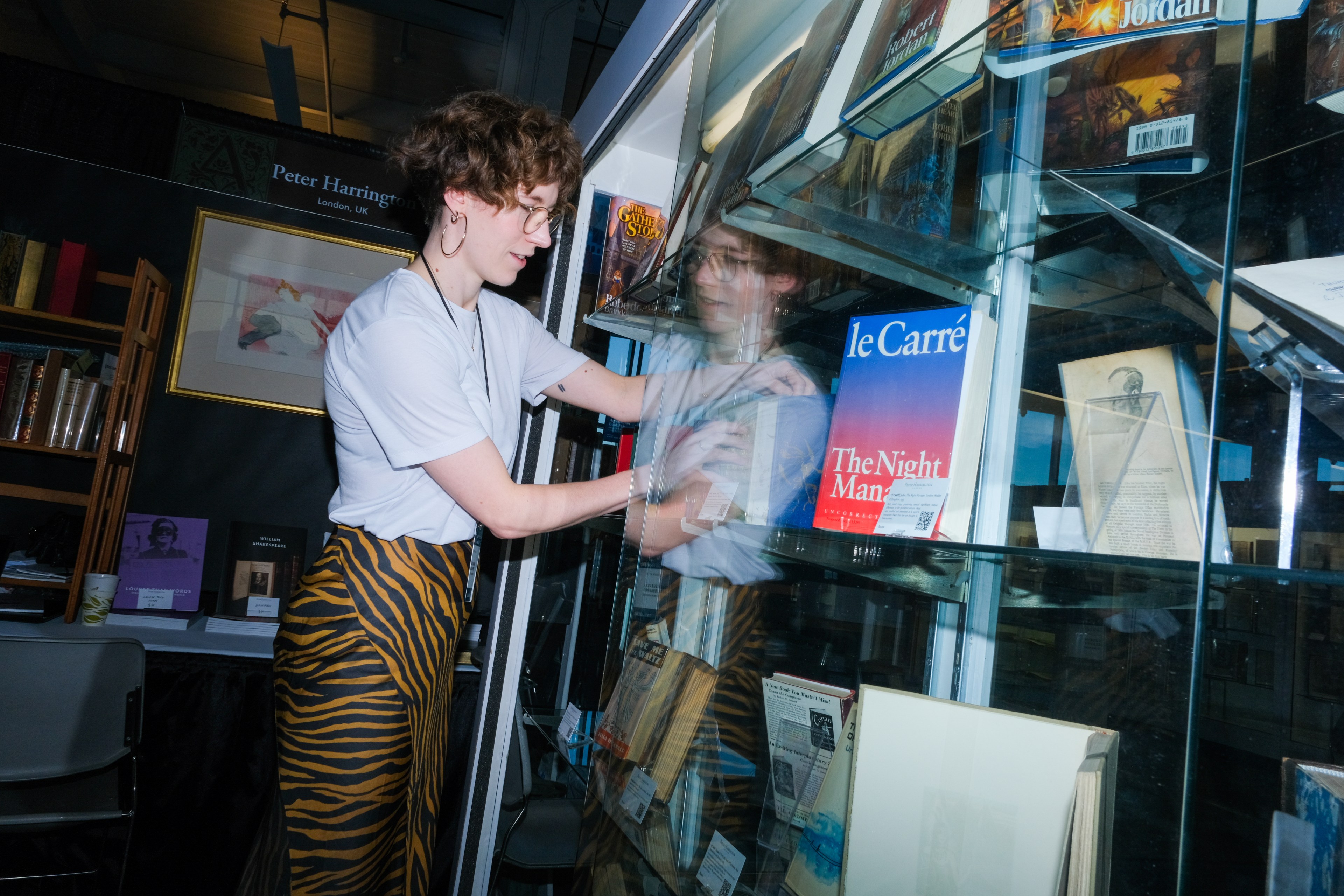 A person in a white shirt and striped pants is arranging books inside a glass display case in a well-lit room.