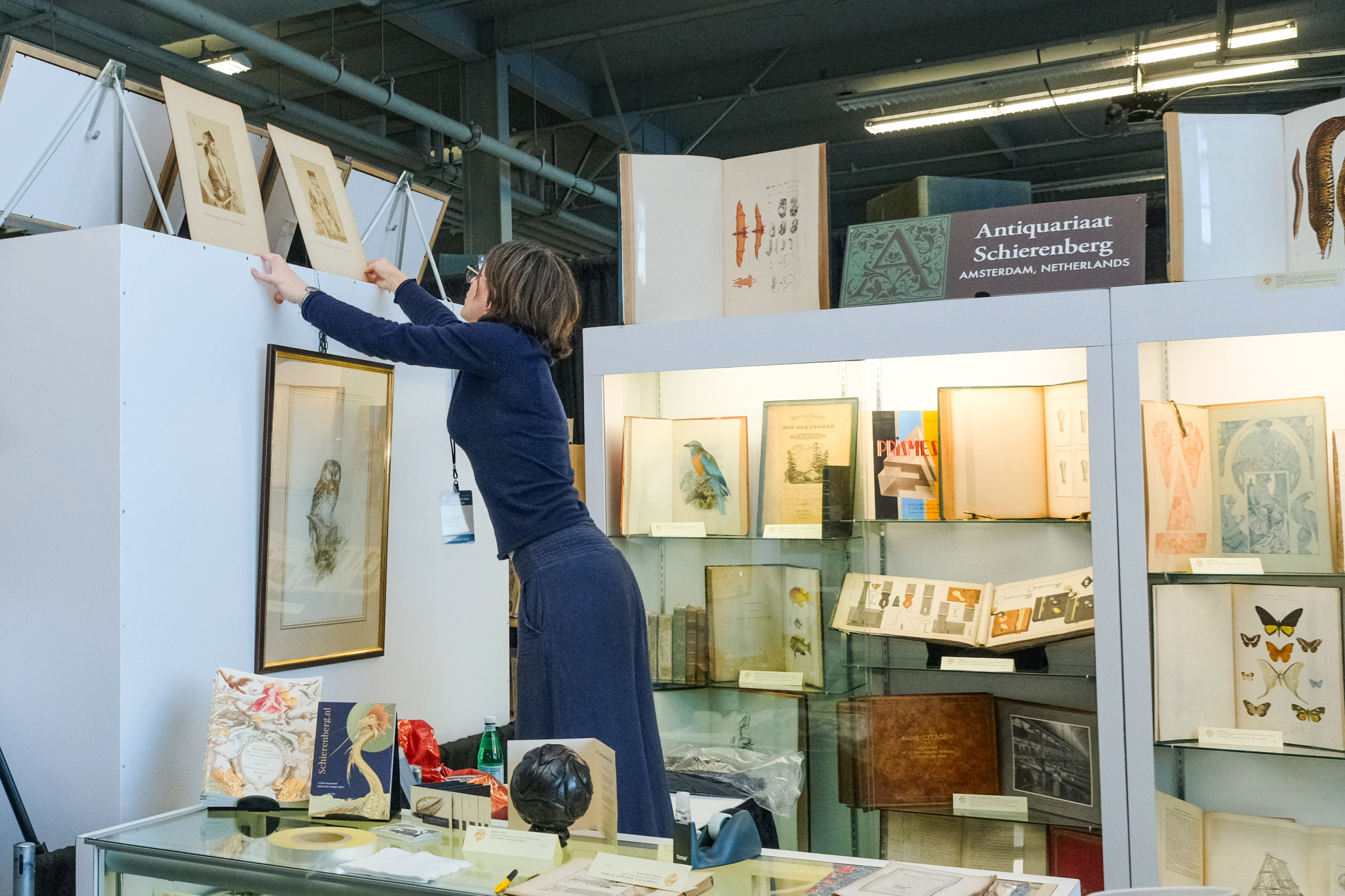A woman is arranging framed artworks on a high shelf at an antique book fair.