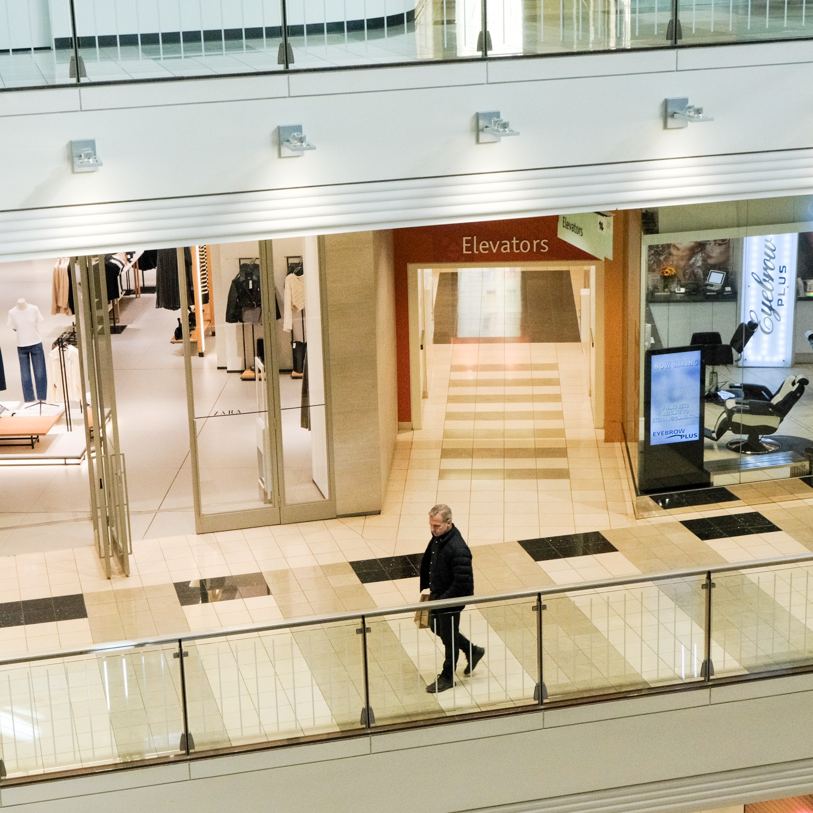 A person walks through a shopping mall with stores on either side. On the left is a clothing store with mannequins, and on the right is an eyebrow salon.