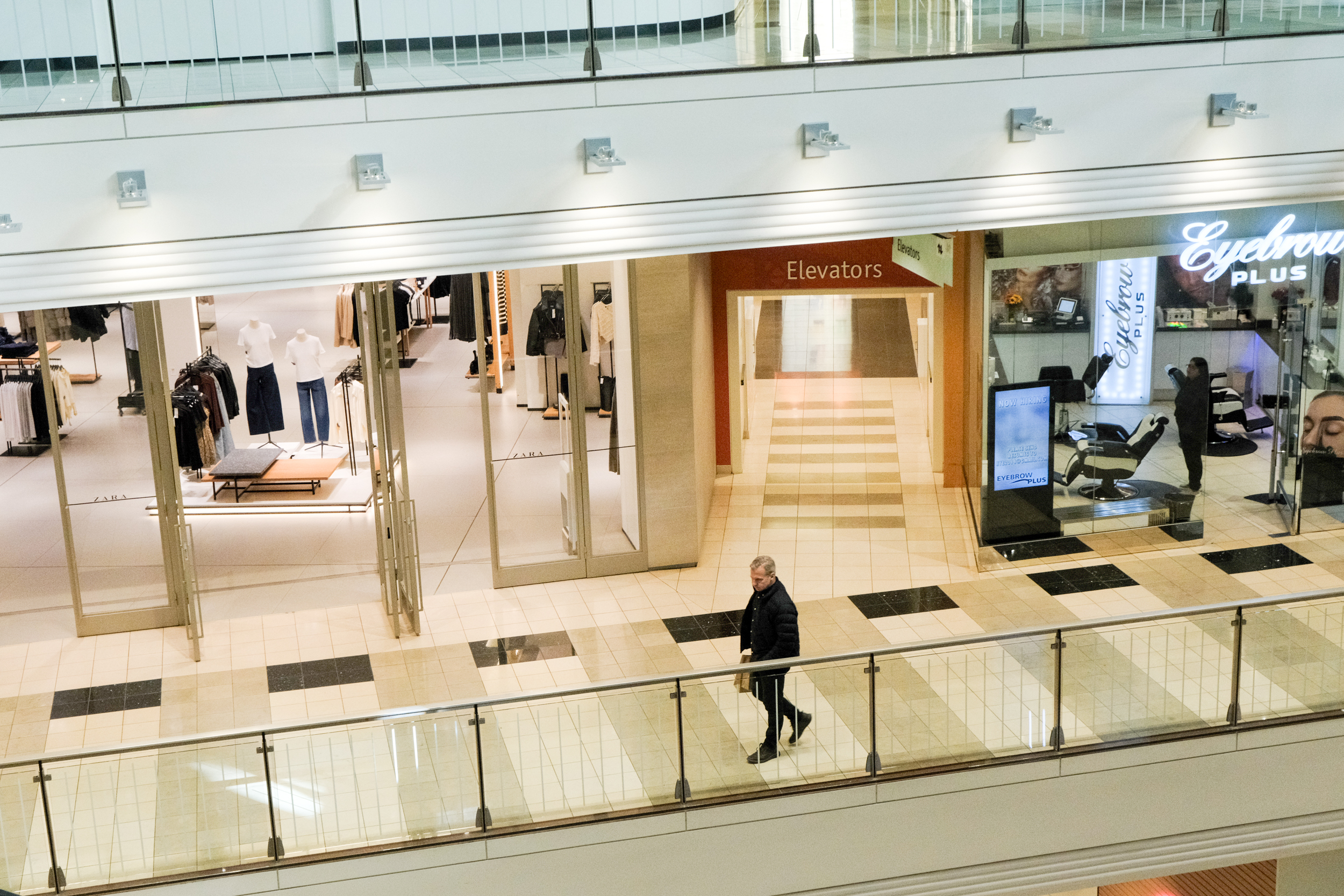 A person walks through a shopping mall with stores on either side. On the left is a clothing store with mannequins, and on the right is an eyebrow salon.