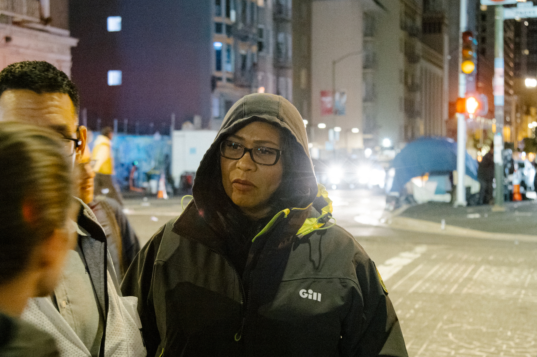 A person in a hooded jacket and glasses stands on a city street at night, with other people and car lights visible.
