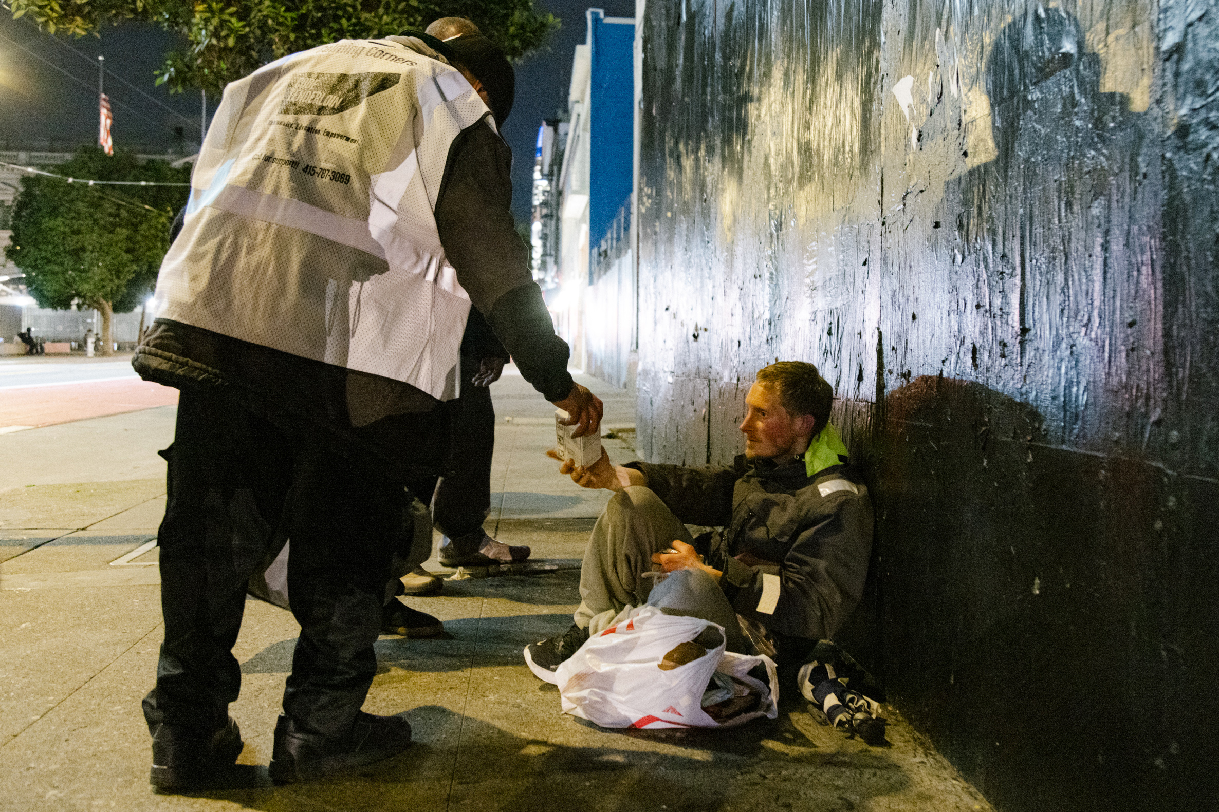 A person in a reflective vest hands a cup to a seated individual against a weathered wall at night.