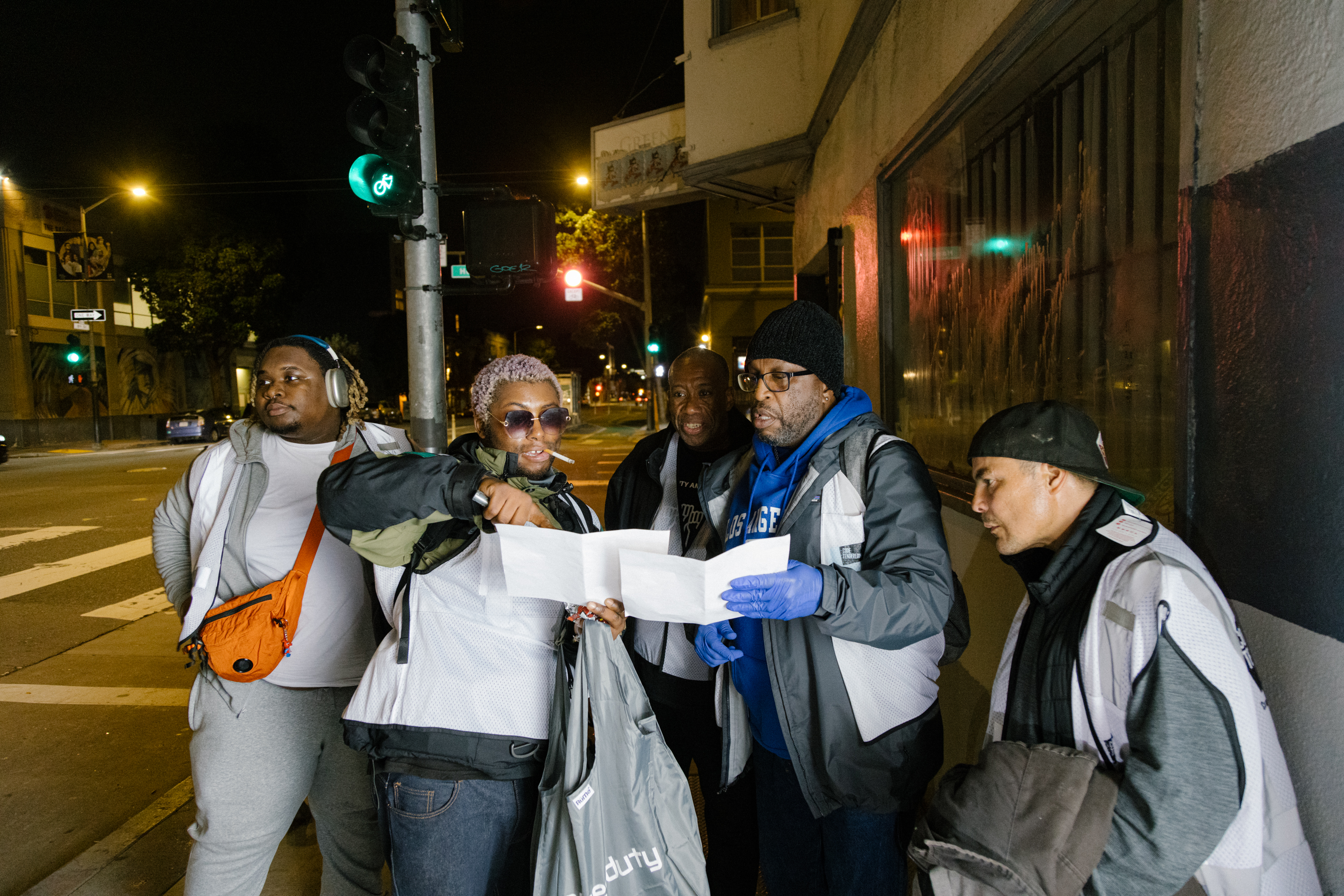 A group of five people stand by a street at night, some focused on reading a paper one person is holding.