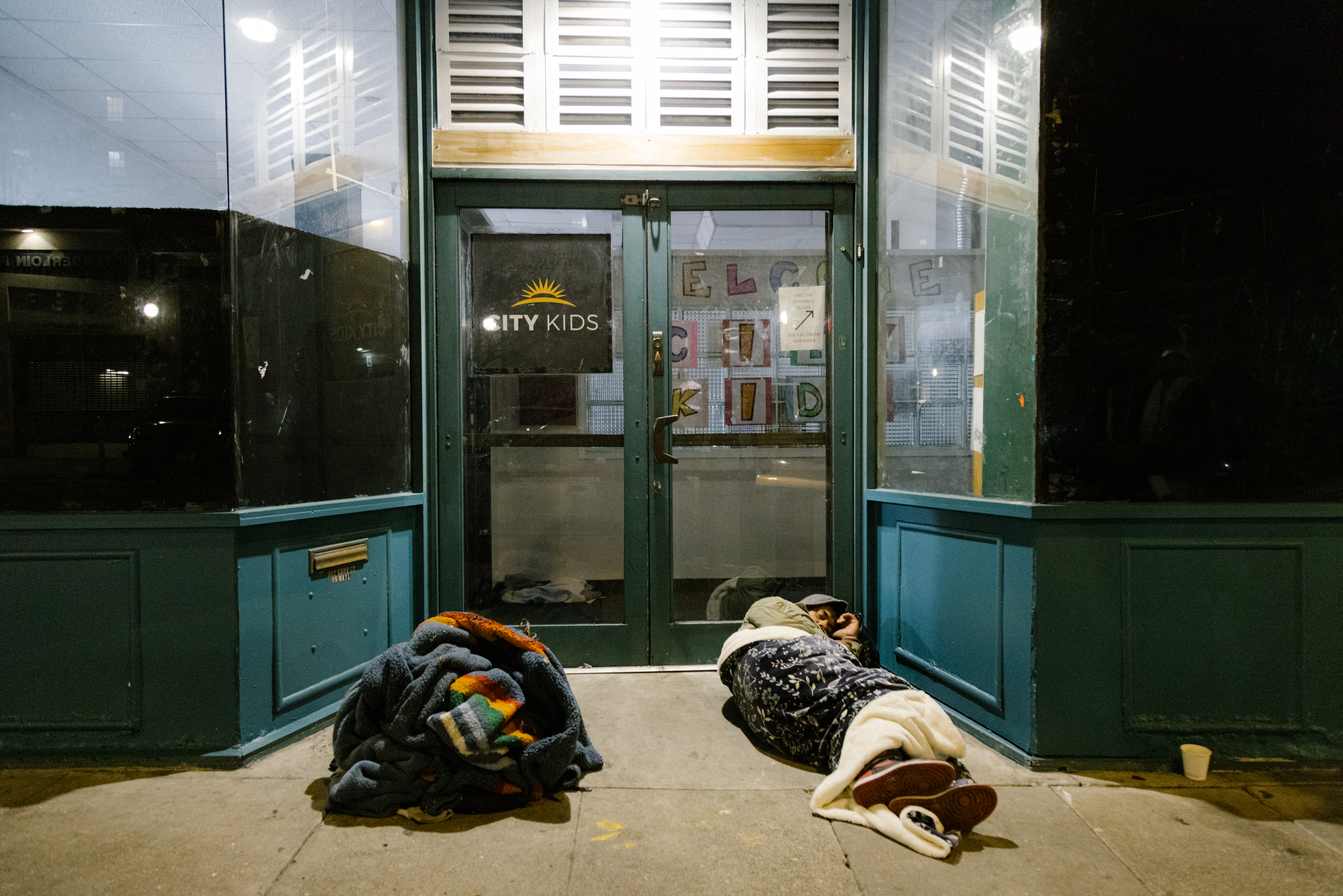 Two people wrapped in blankets lay on the sidewalk at night in front of a storefront