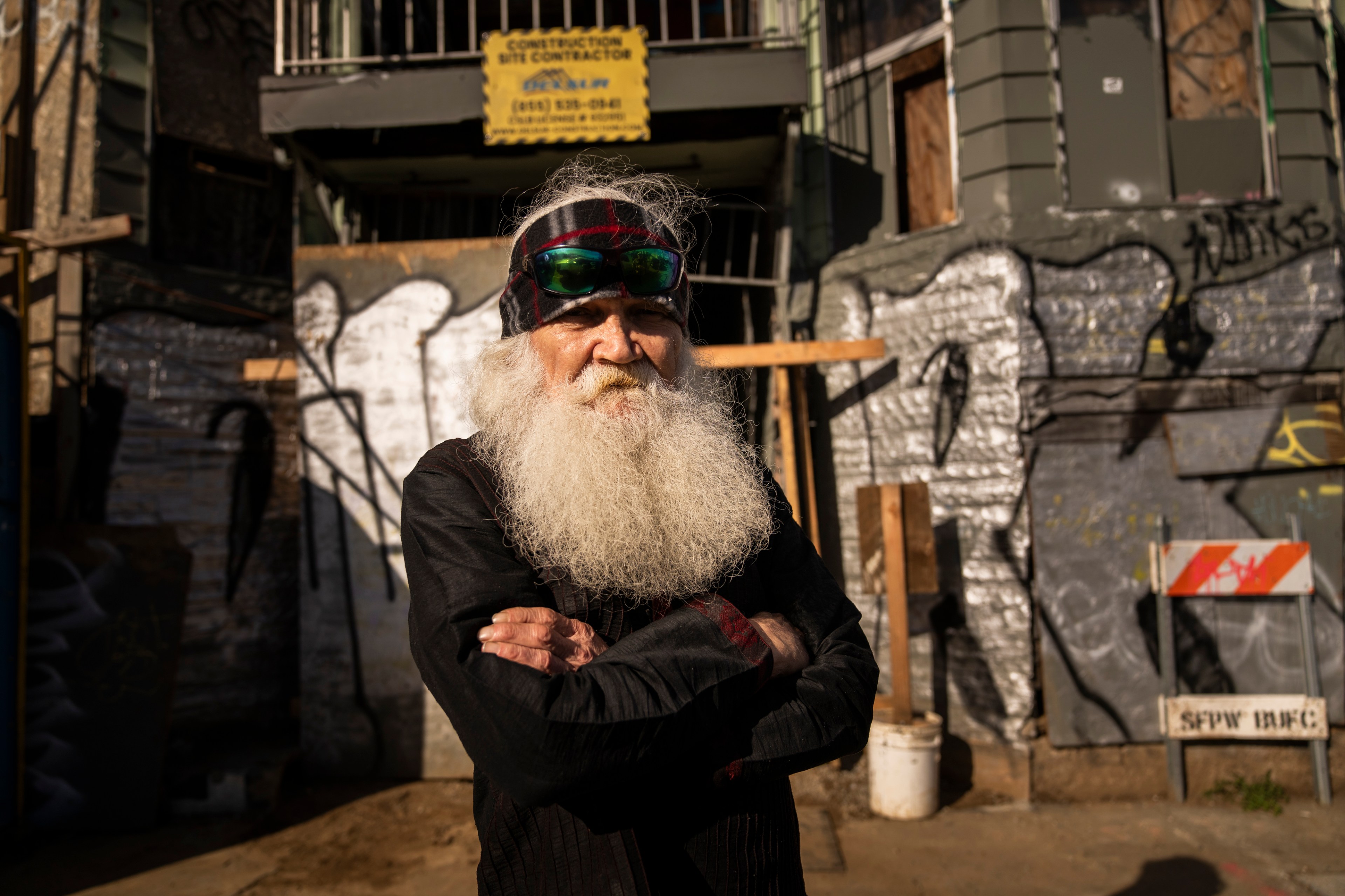 An elder man with a voluminous white beard, wearing sunglasses and a bandana, stands confidently with crossed arms against an urban backdrop with graffiti.