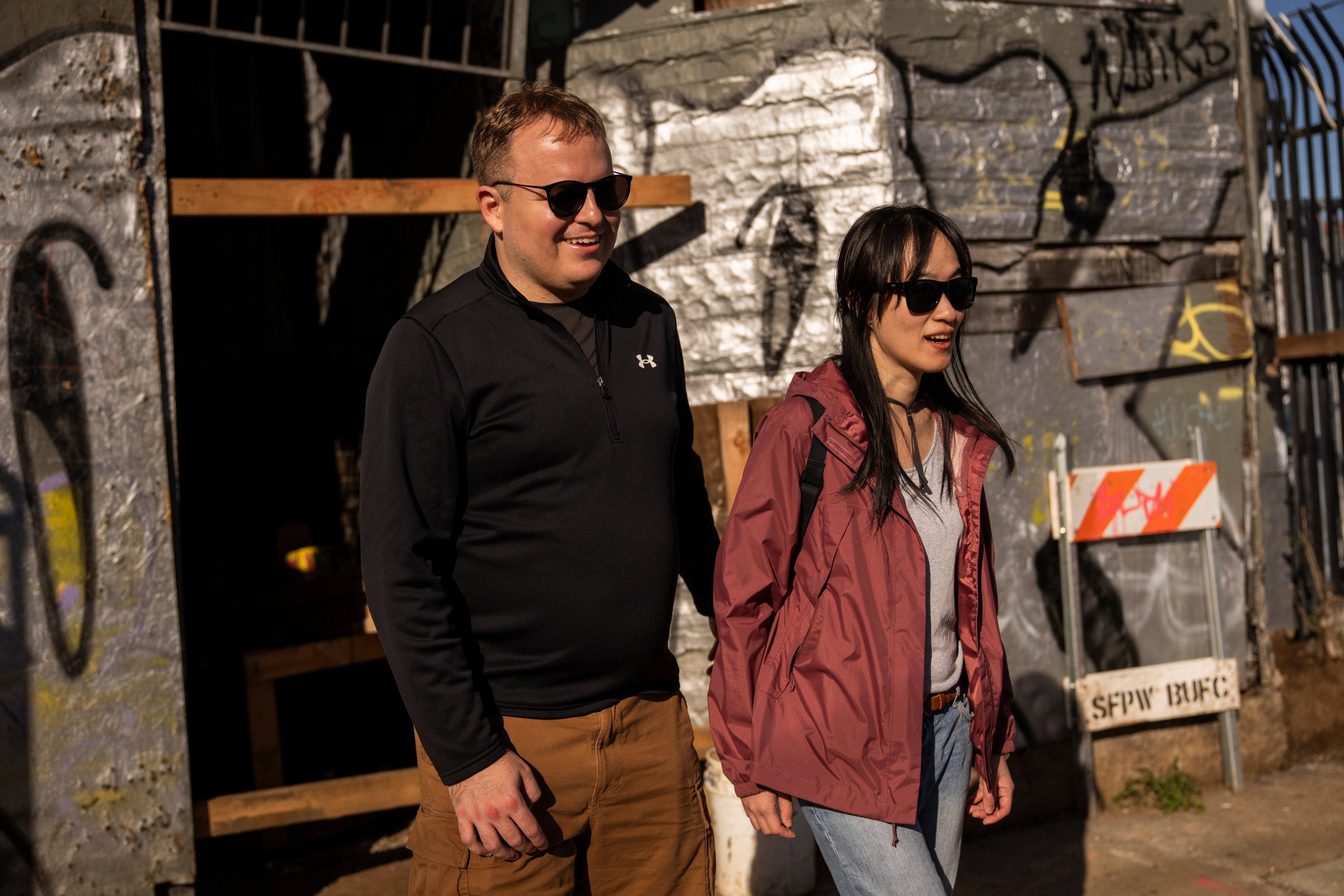 A man and a woman, both wearing sunglasses, walk by graffiti-covered walls in sunlight.