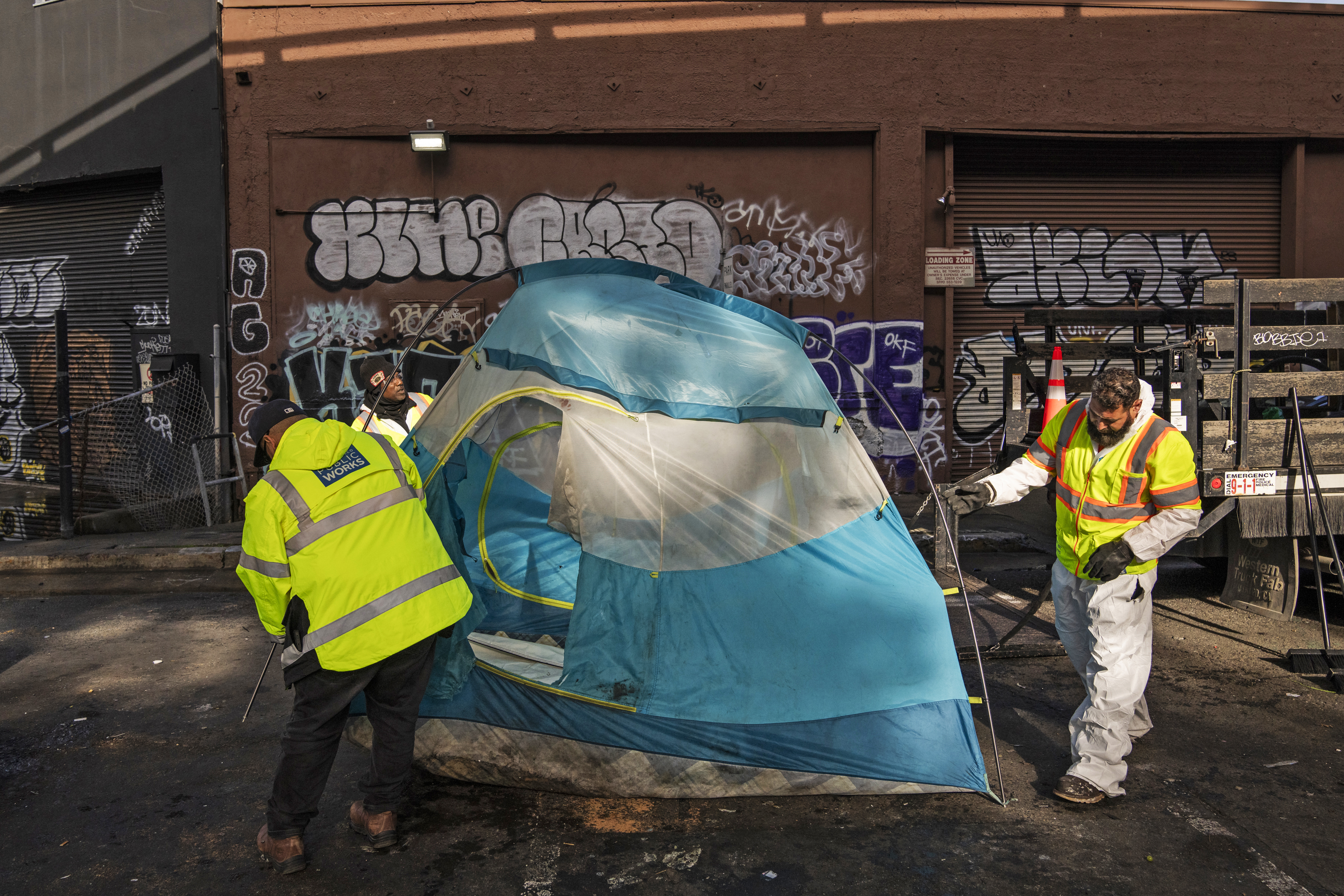 Workers in high-visibility vests handle a blue tent on an urban street with graffiti-covered walls.
