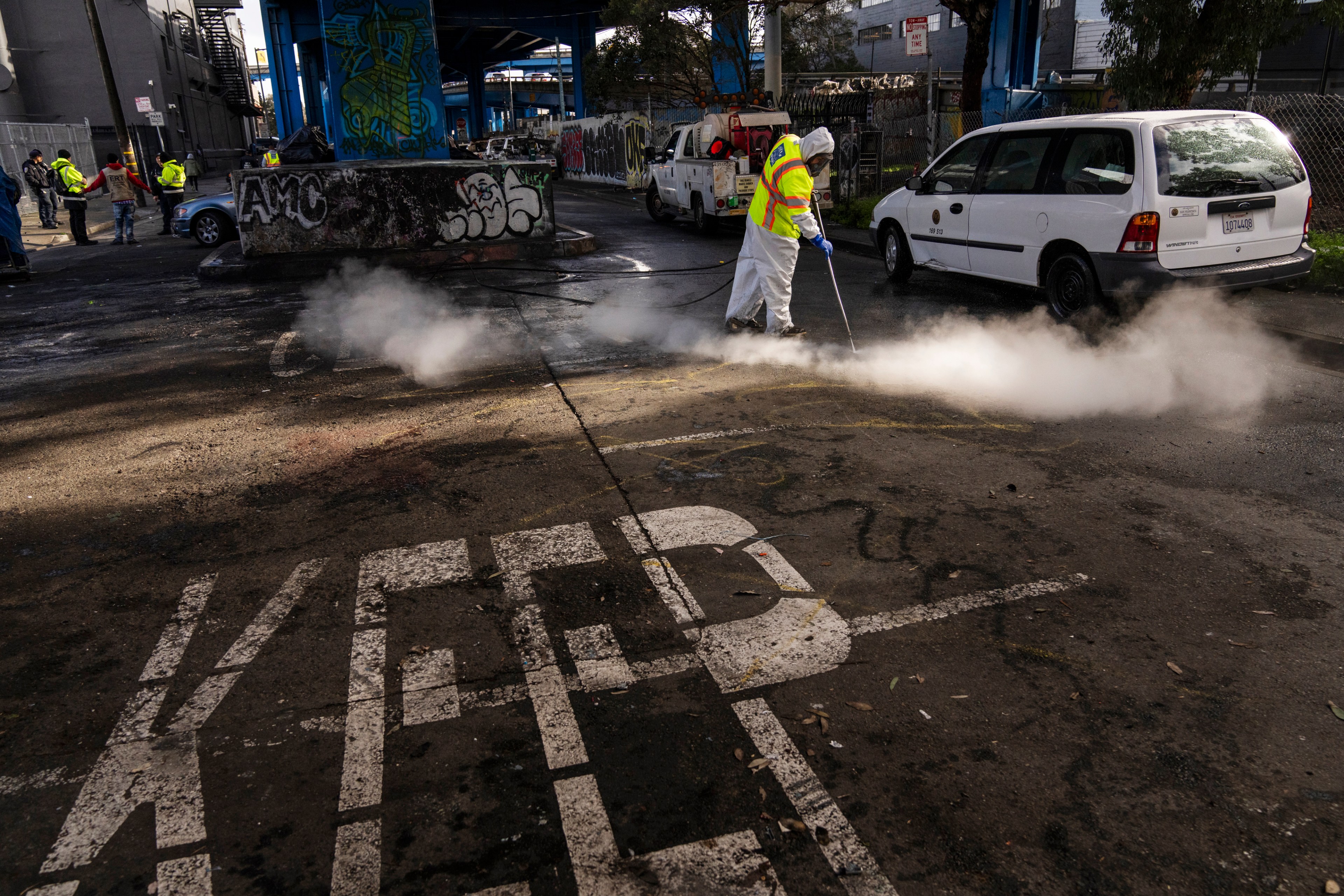 A worker in protective gear pressure-washes a street emitting steam, near graffiti-covered walls and parked cars, while onlookers, some in reflective vests, observe.