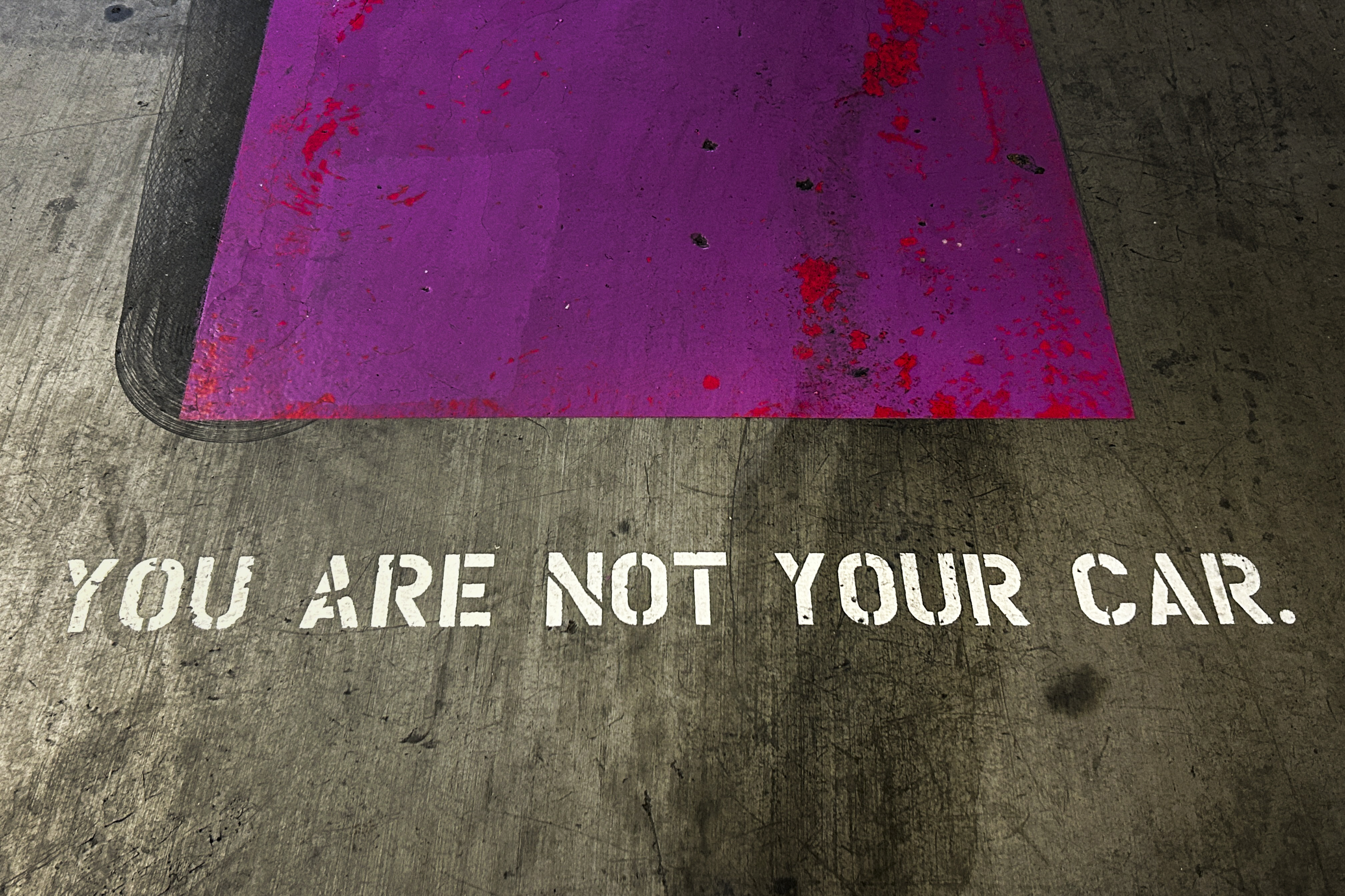 A purple parking spot with red splatters on a gray floor has &quot;YOU ARE NOT YOUR CAR&quot; stenciled in white.