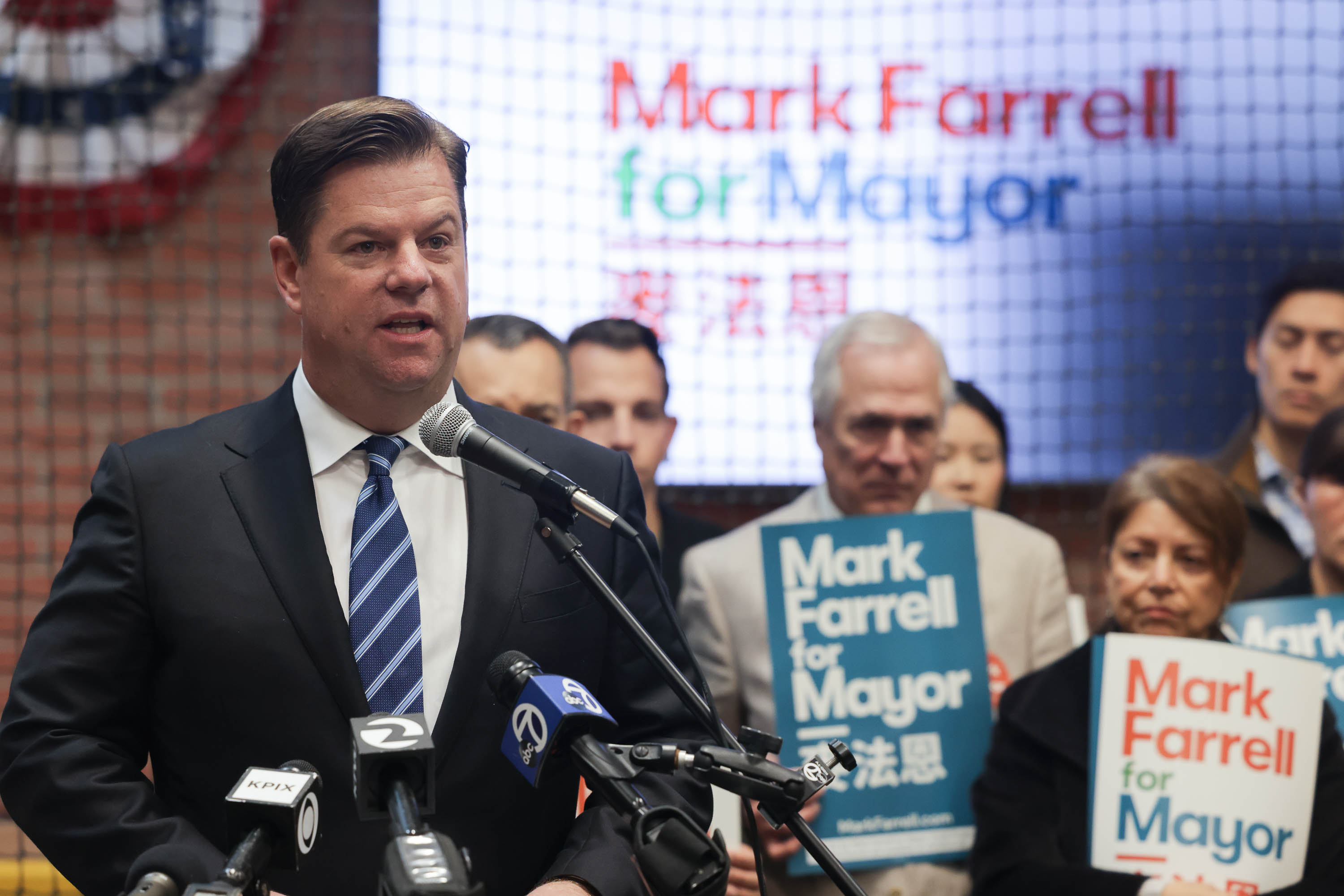A man speaks in front of microphones as supporters holding campaign signs reading &quot;Mark Farrell for mayor&quot; stand in the background.