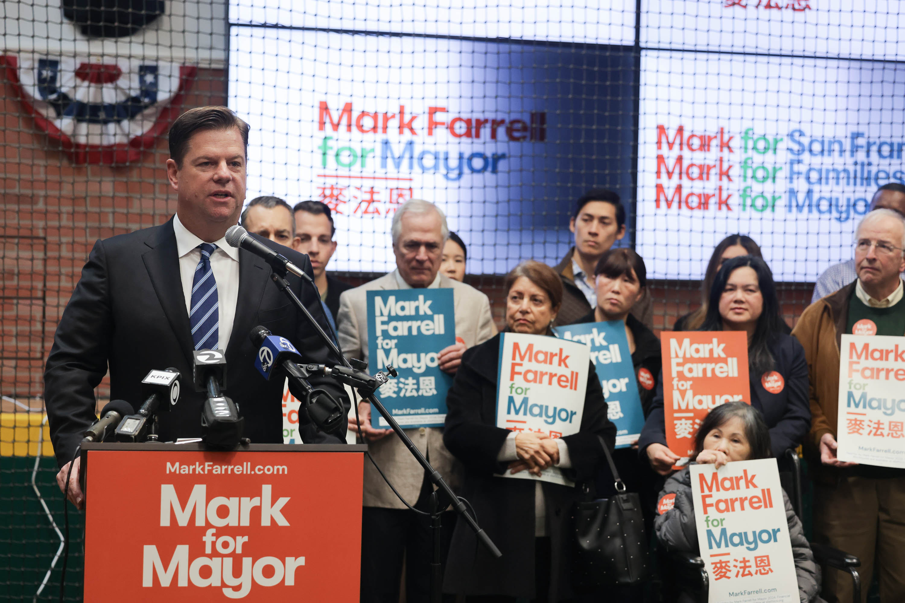 A man stands at a podium as a crowd stands in the background holding &quot;Mark Farrell for mayor&quot; signs. 