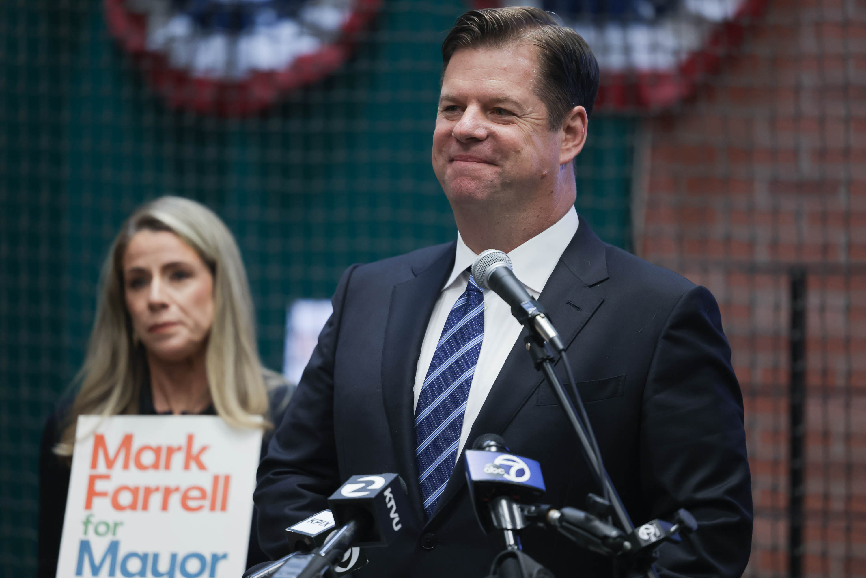 Mark Farrell, wearing a suit and tie, smiles at a podium with microphones as his wife holds &quot;Mark Farrell for Mayor&quot; sign behind him.