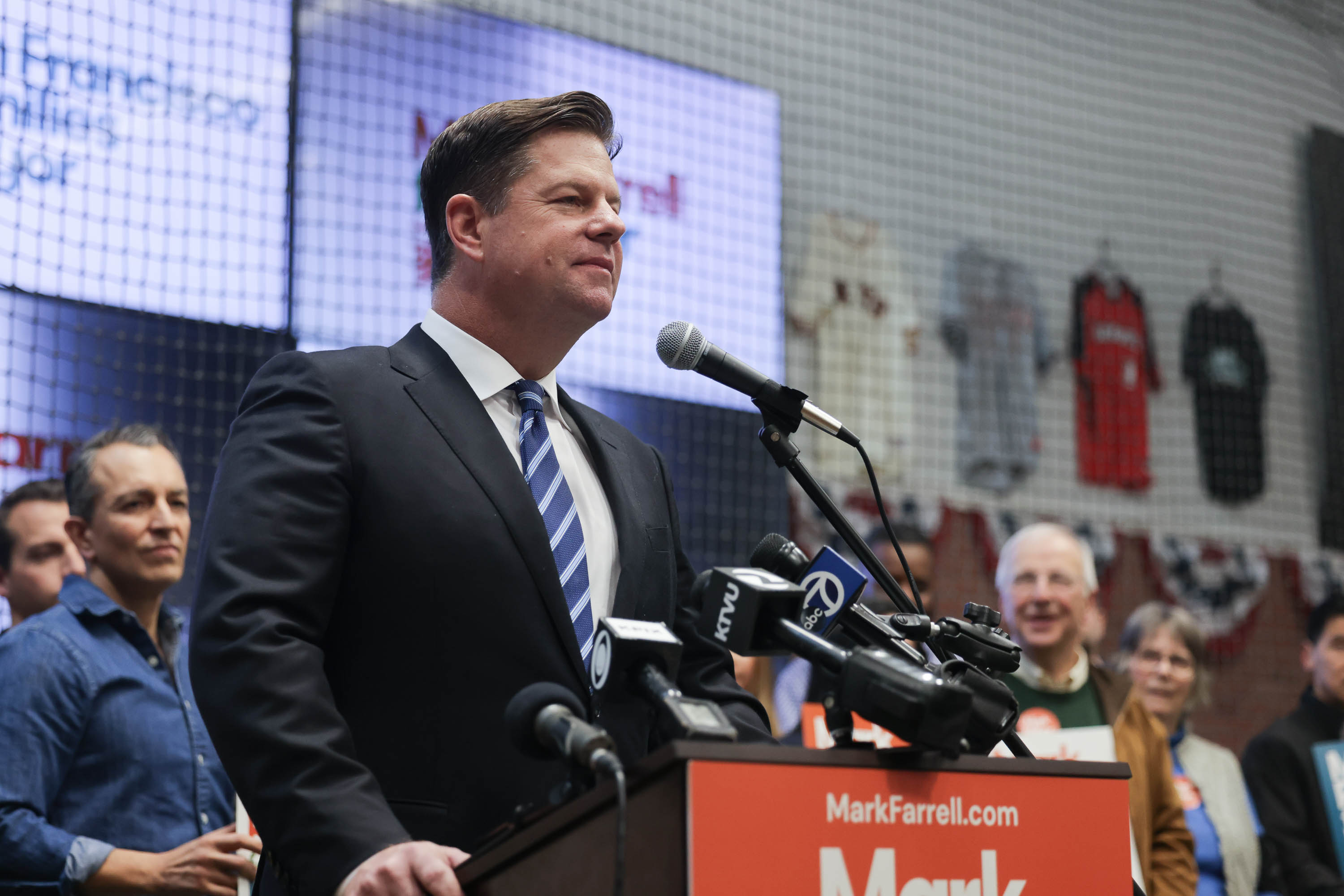 Mark Farrell in a suit speaks at a podium with microphones, a campaign banner in front, and onlookers behind him.