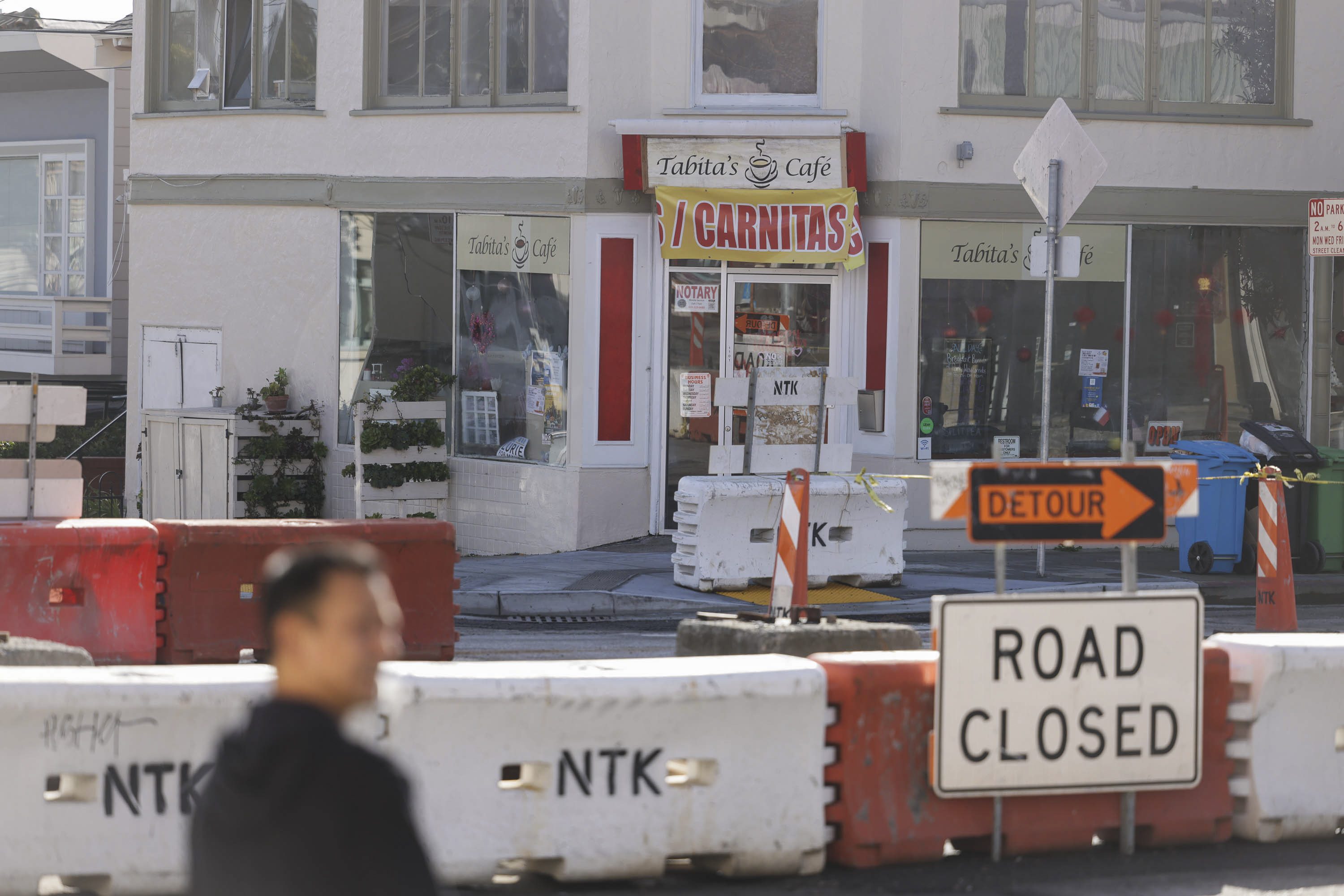 A cafe behind road barriers with &quot;ROAD CLOSED&quot; and &quot;DETOUR&quot; signs, with a blurred person in the foreground.