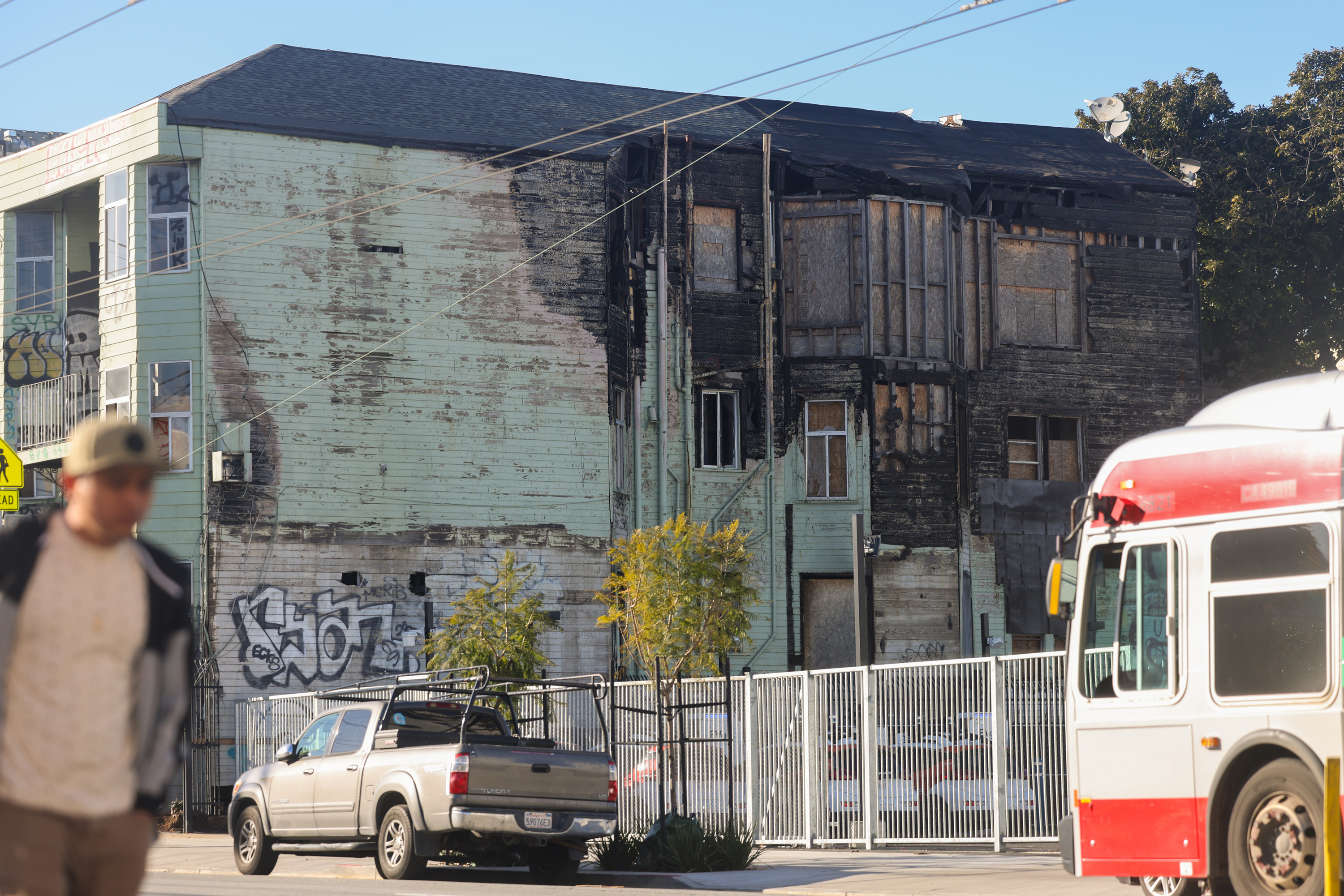 A dilapidated two-story building with charred walls, graffiti, and a fence in an urban setting, with a blurred pedestrian and bus in the foreground.