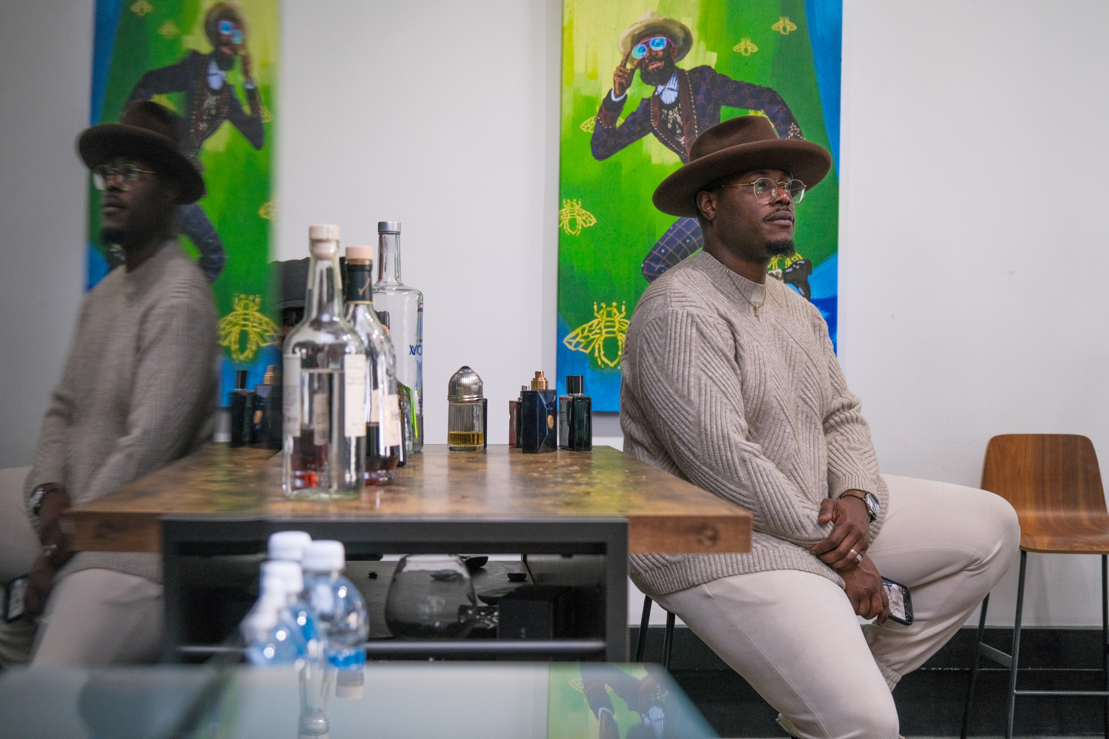 A man in a hat sits near a table with bottles, his reflection visible, and vibrant art hangs behind.