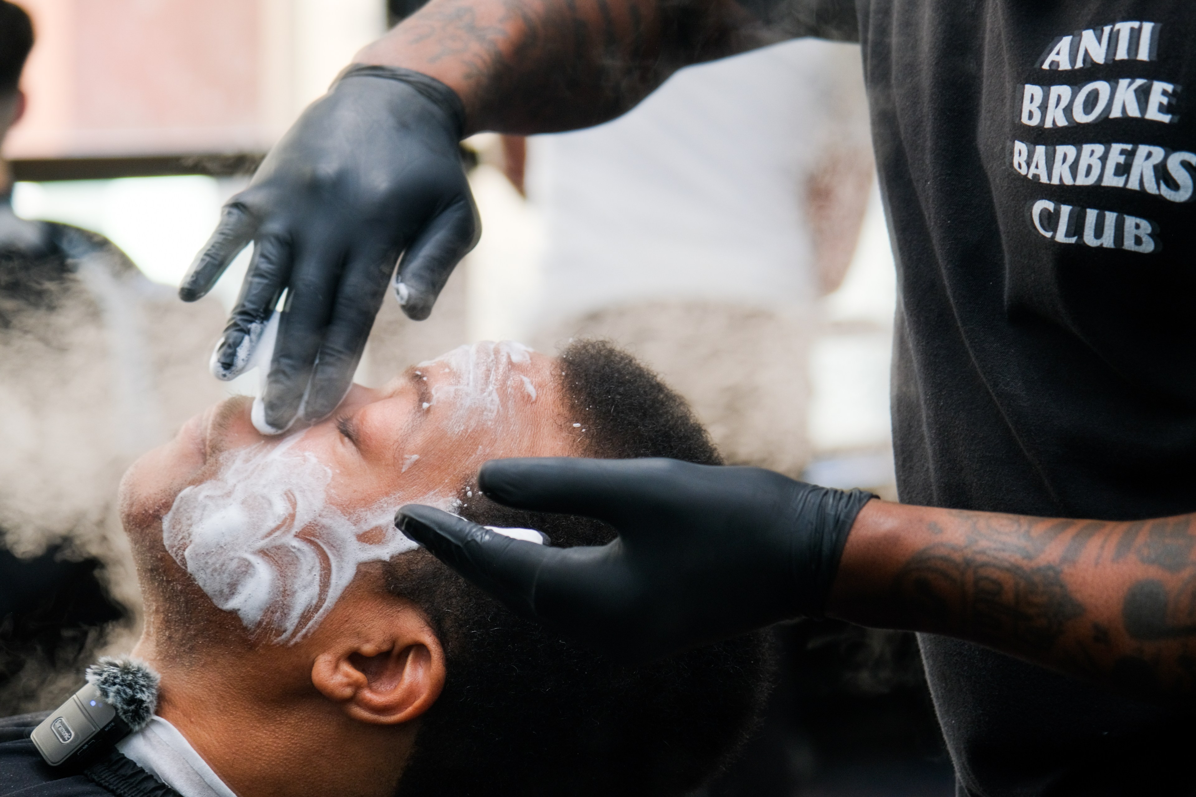 A barber wearing gloves applies shaving cream to a reclined customer's face.
