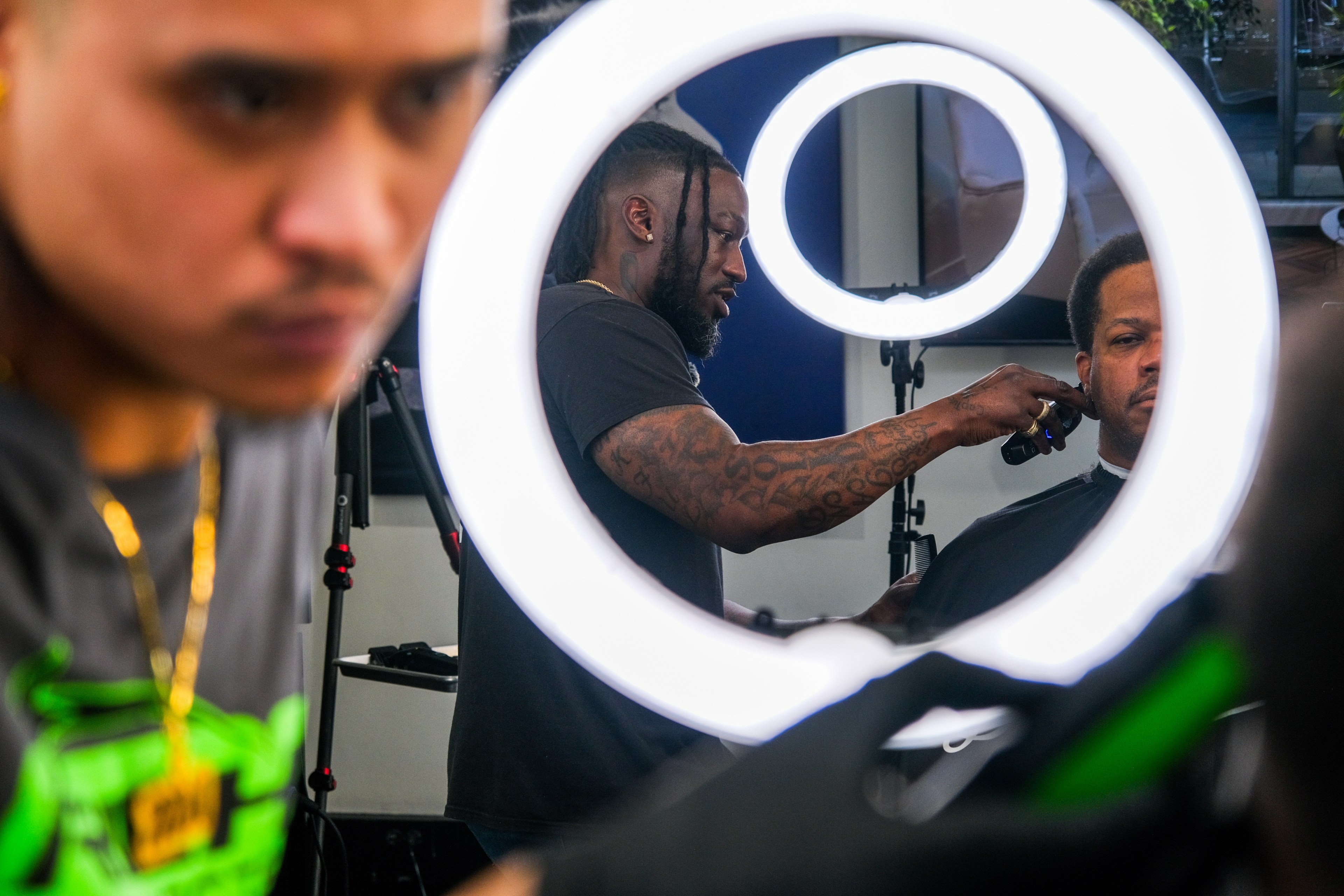A barber with tattoos gives a haircut, framed by a ring light; another person in foreground, out of focus.
