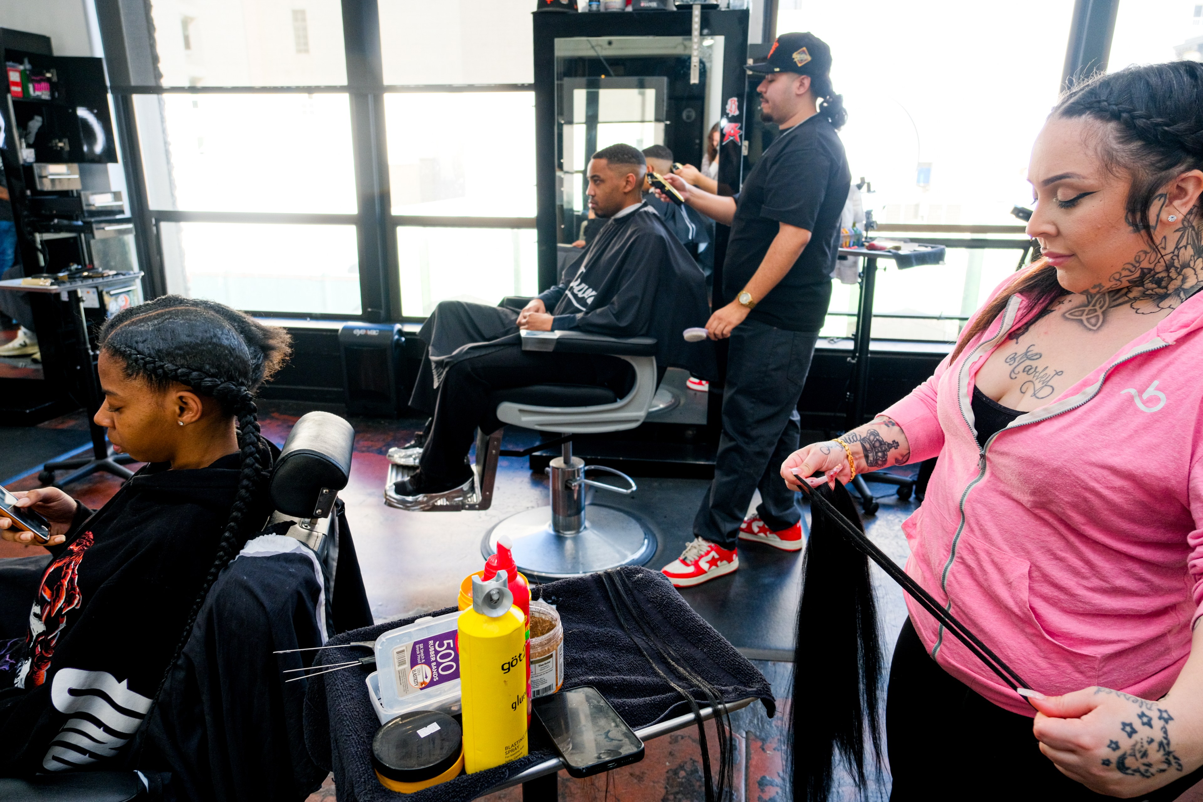Inside a brightly lit barbershop, two barbers style clients' hair, with hair care products in the foreground.