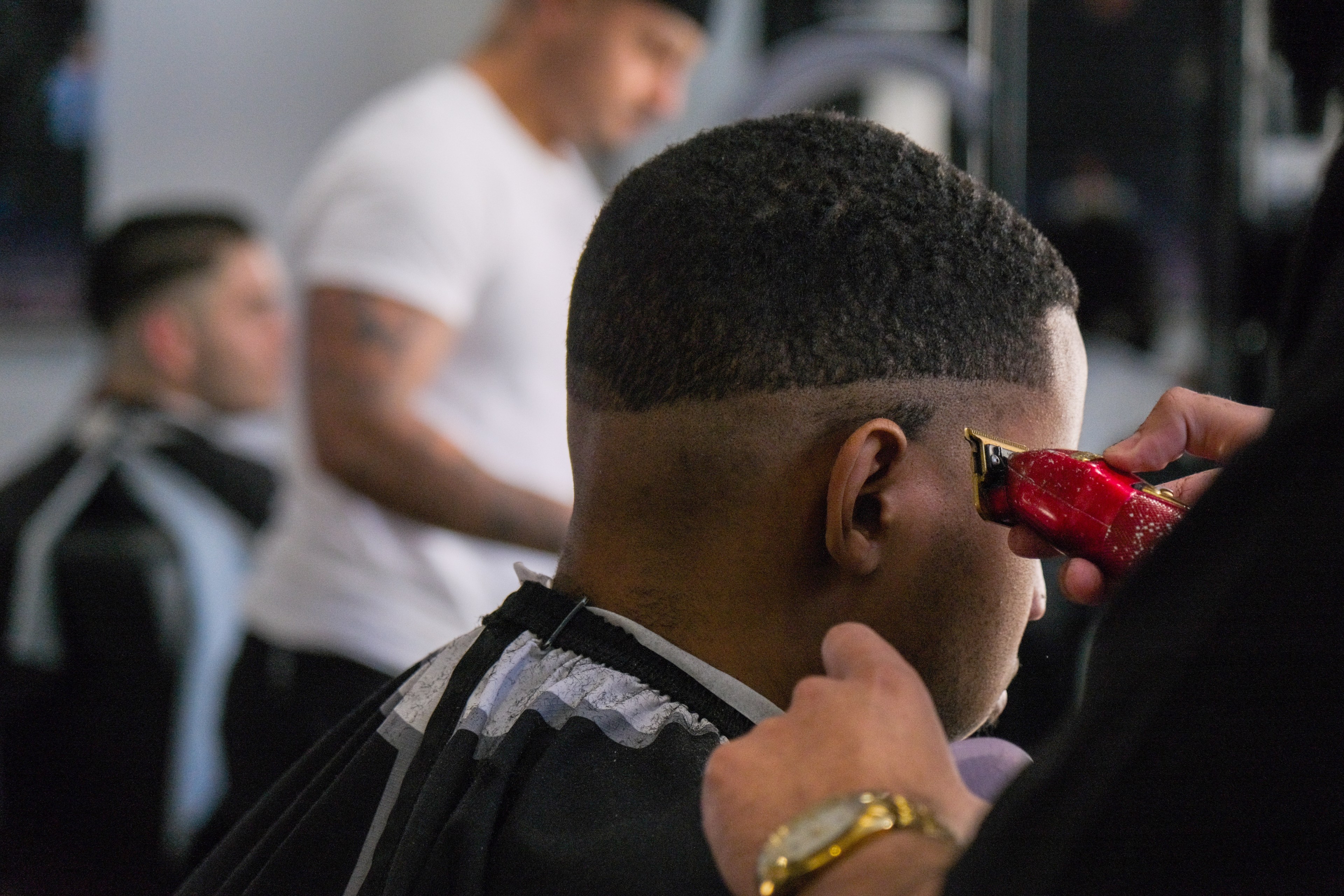 A barber uses red clippers to trim a man's hairline; another customer and barber are blurry in the background.