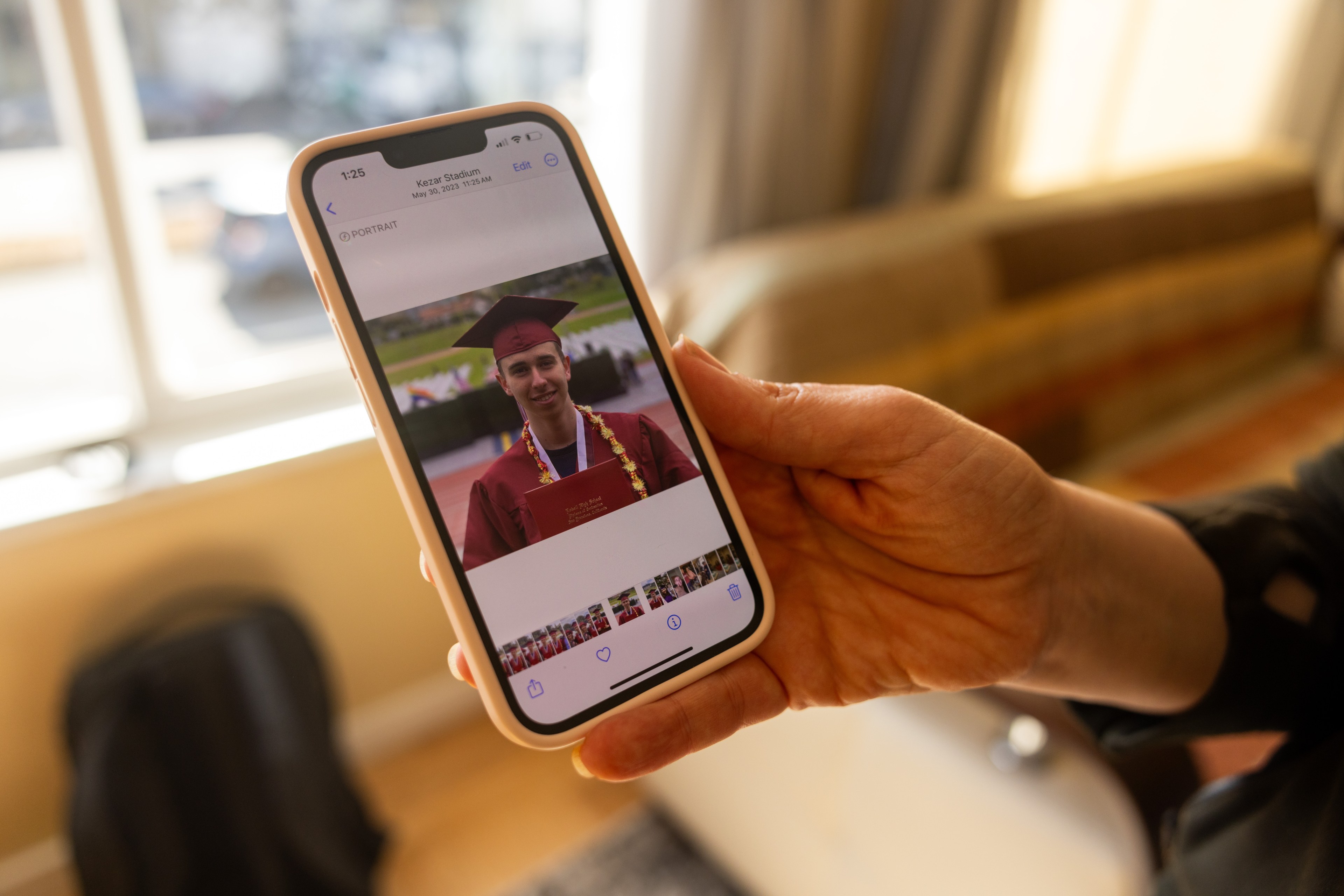 A person holds a phone displaying a photo of a graduate in a maroon cap and gown. The image shows the graduate smiling, holding a diploma.