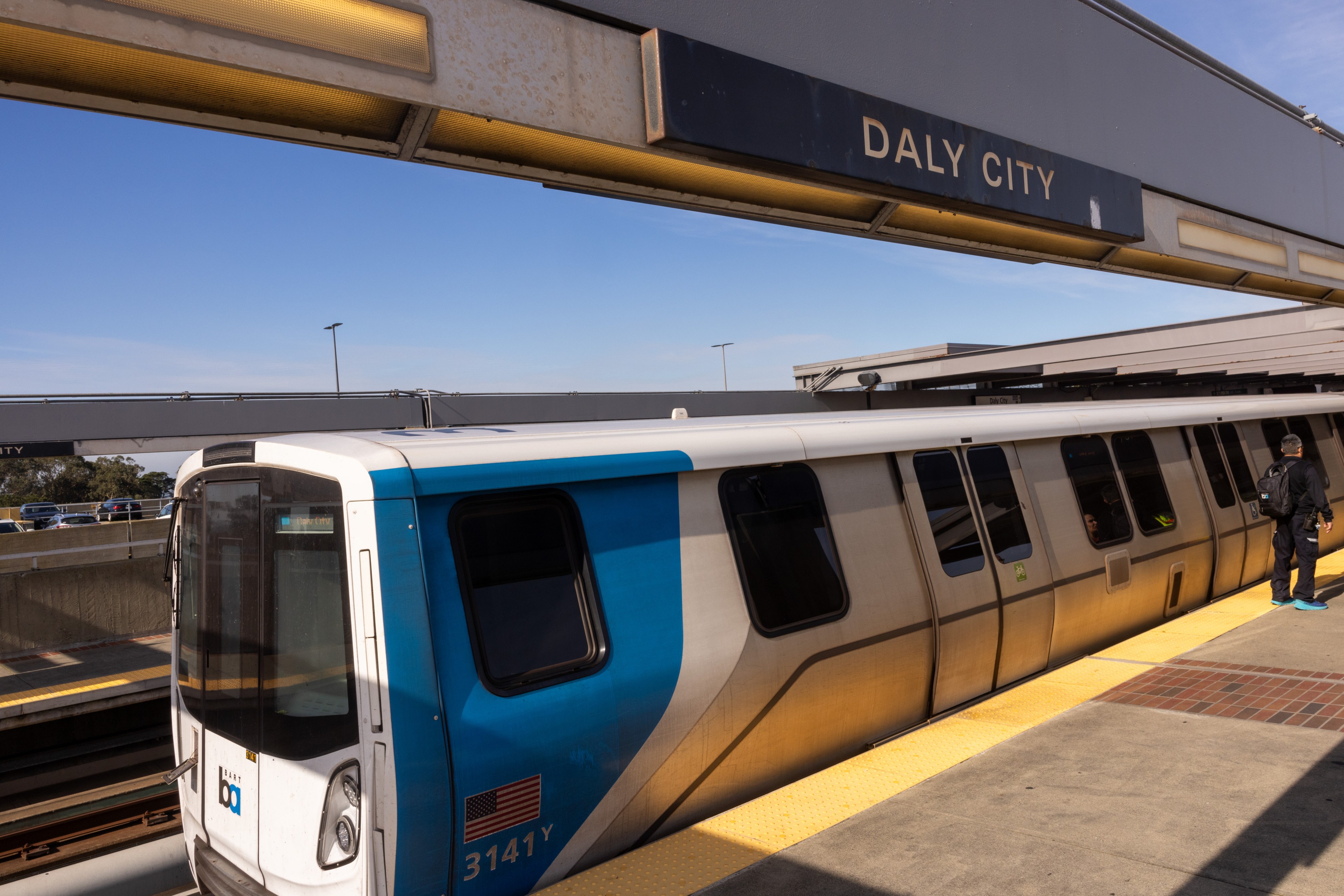A train at Daly City station with a person on the platform under a clear sky.