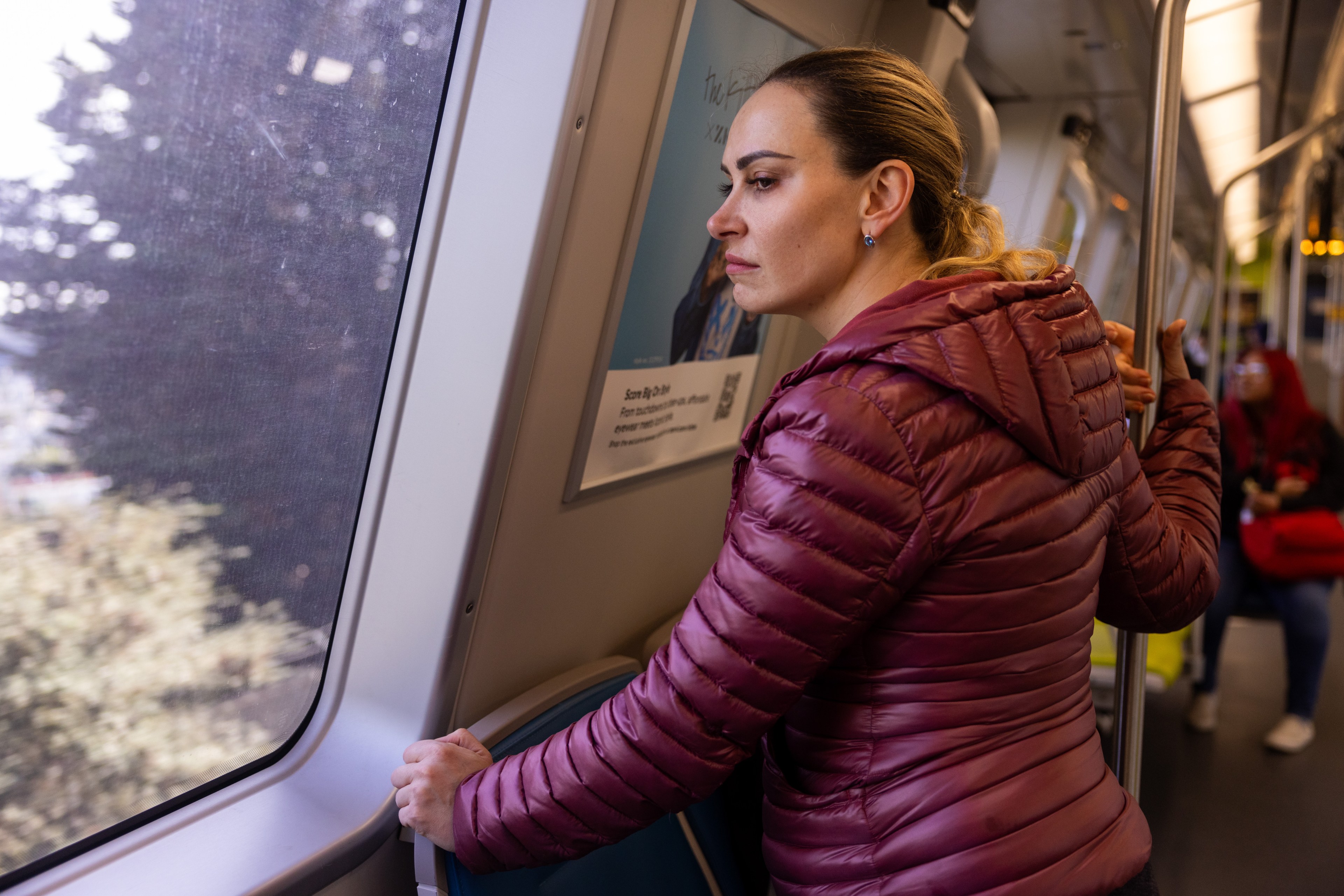 A woman gazes out a train window, trees blur past. She wears a maroon jacket, holding onto a rail, seemingly lost in thought.