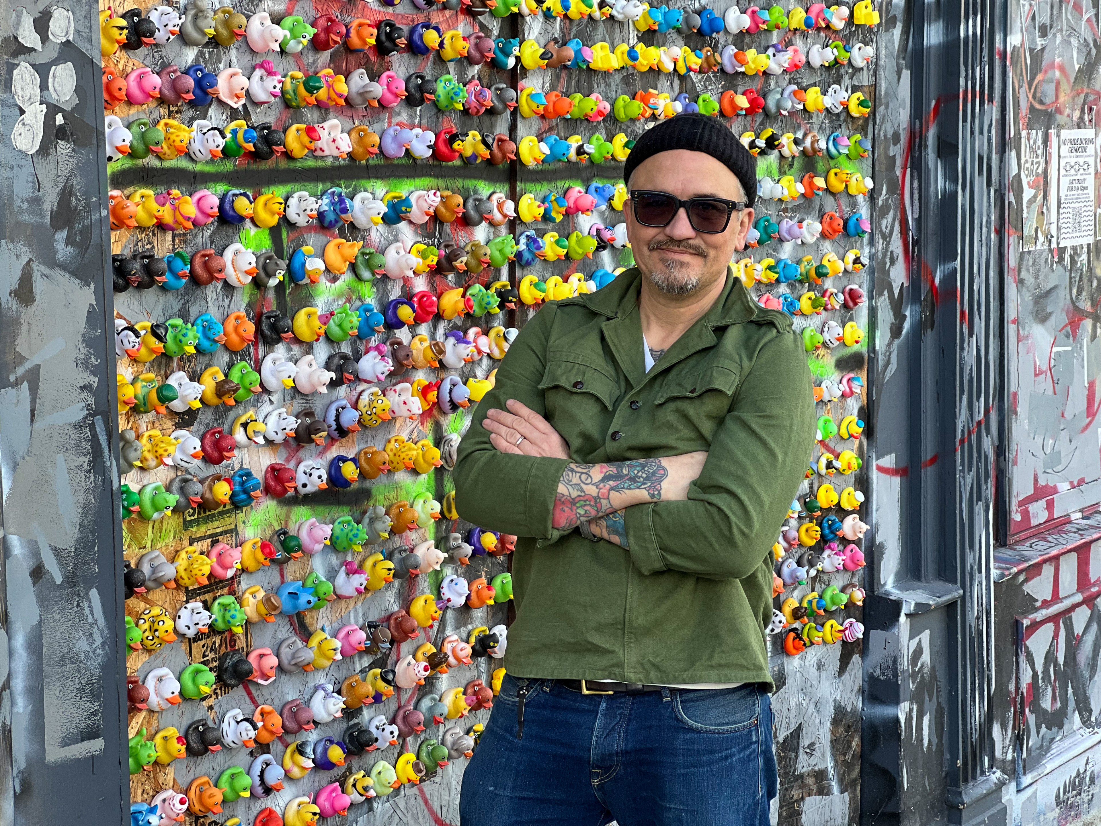 A man stands arms crossed before a vibrant wall of colorful rubber ducks.