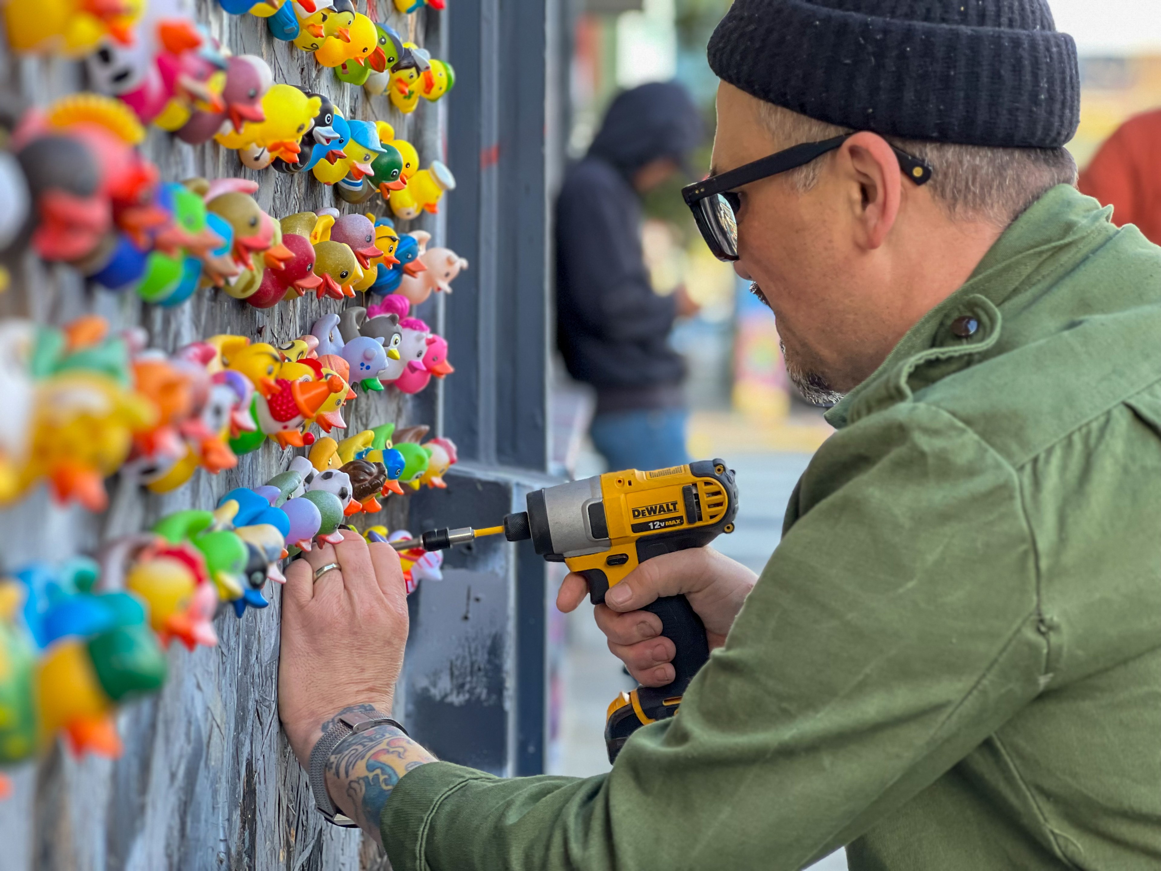 a man in a green shirt and black cap drills a duck into a wall of rubber ducks
