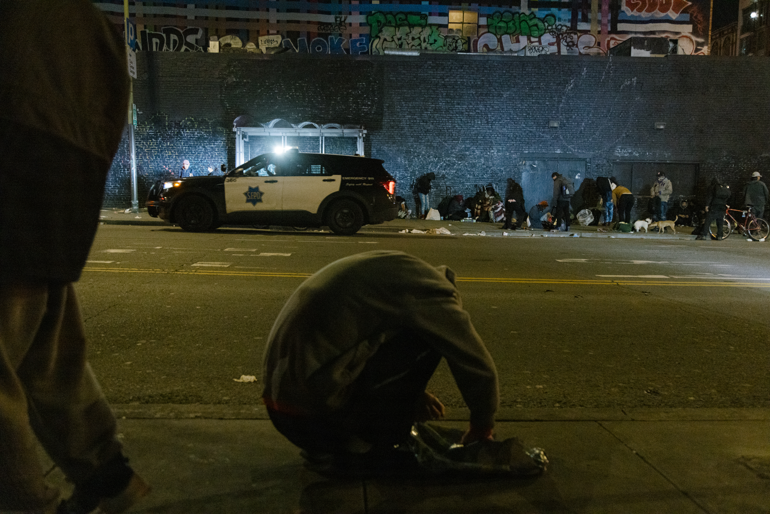 A person crouches on the side of the street as a police car drives by a group of people standing on the sidewalk.