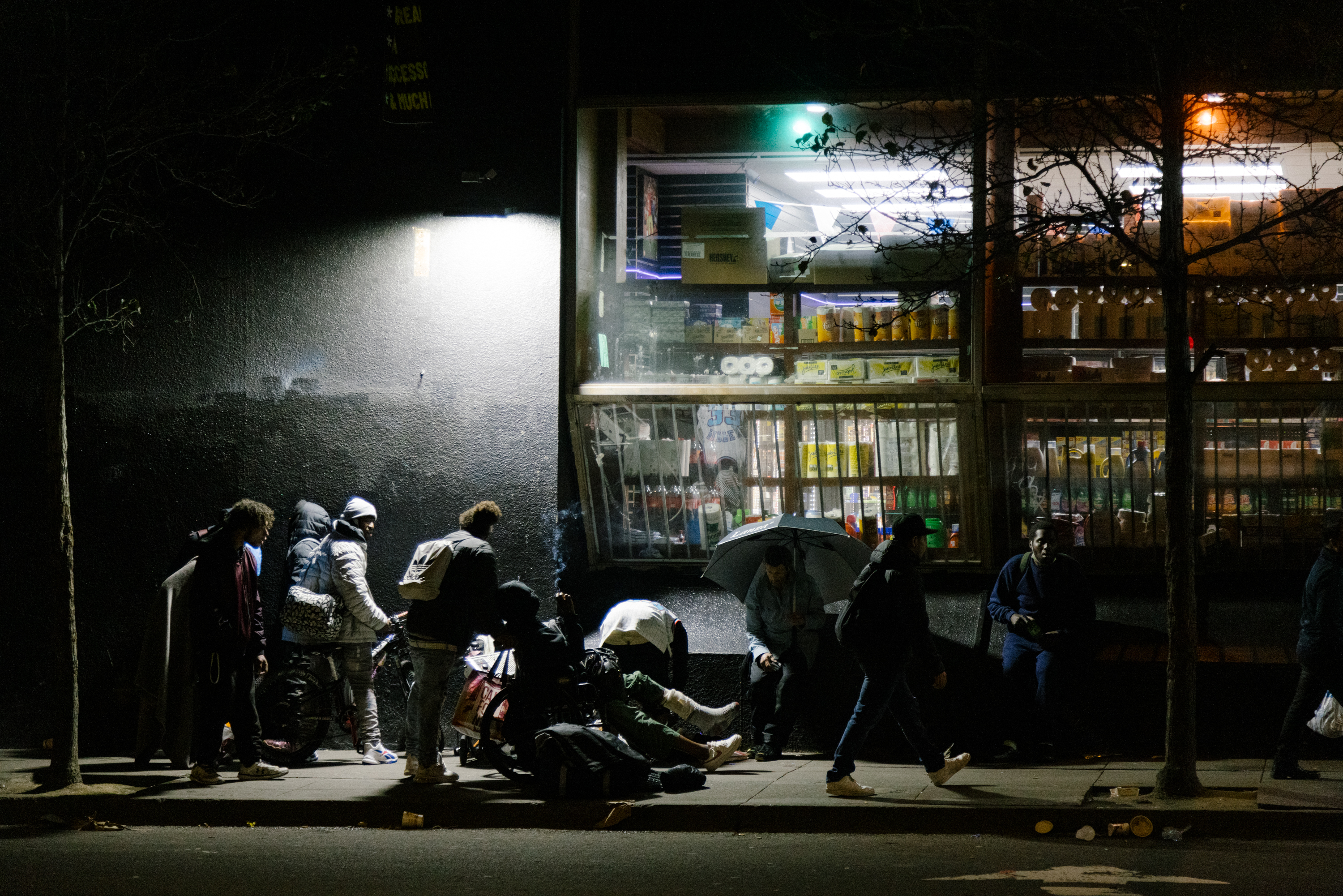 A group gathers at night outside a brightly lit shop, with some on foot and others on bikes.