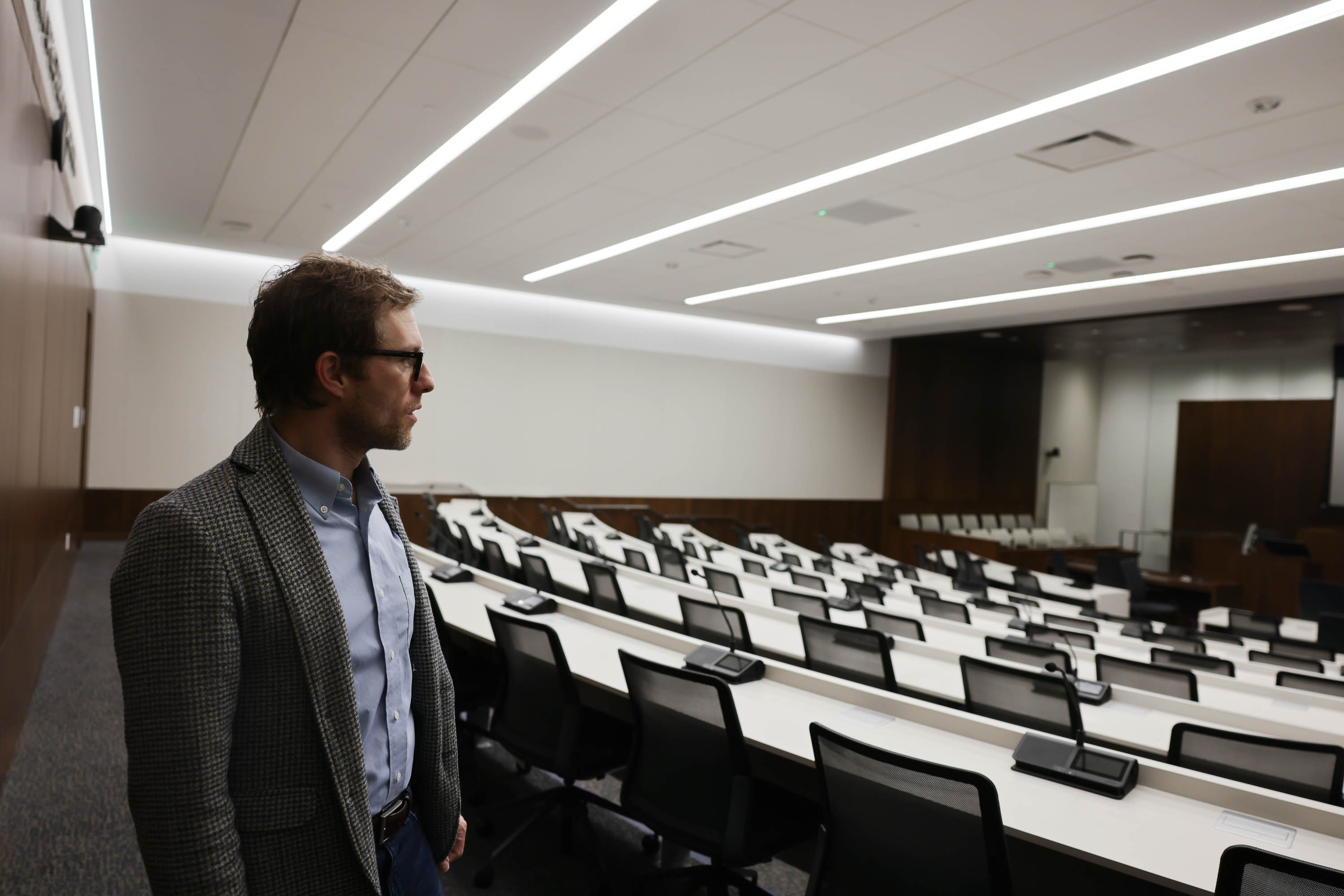 A man in a blazer stands in an empty, modern lecture hall with rows of seats and desks.