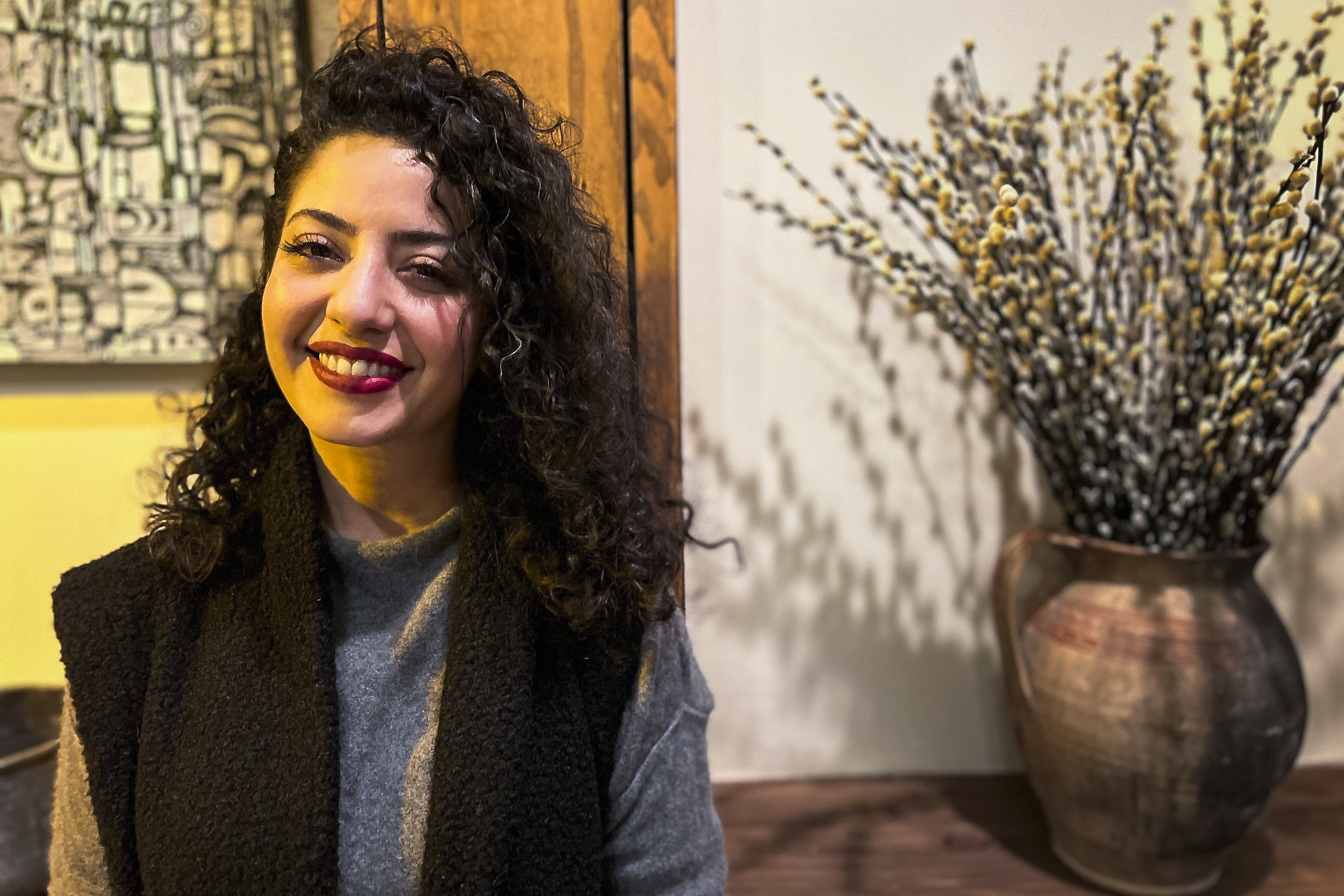 A smiling woman with curly hair stands next to a vase of willow catkins on a wooden surface, with abstract art in the background.