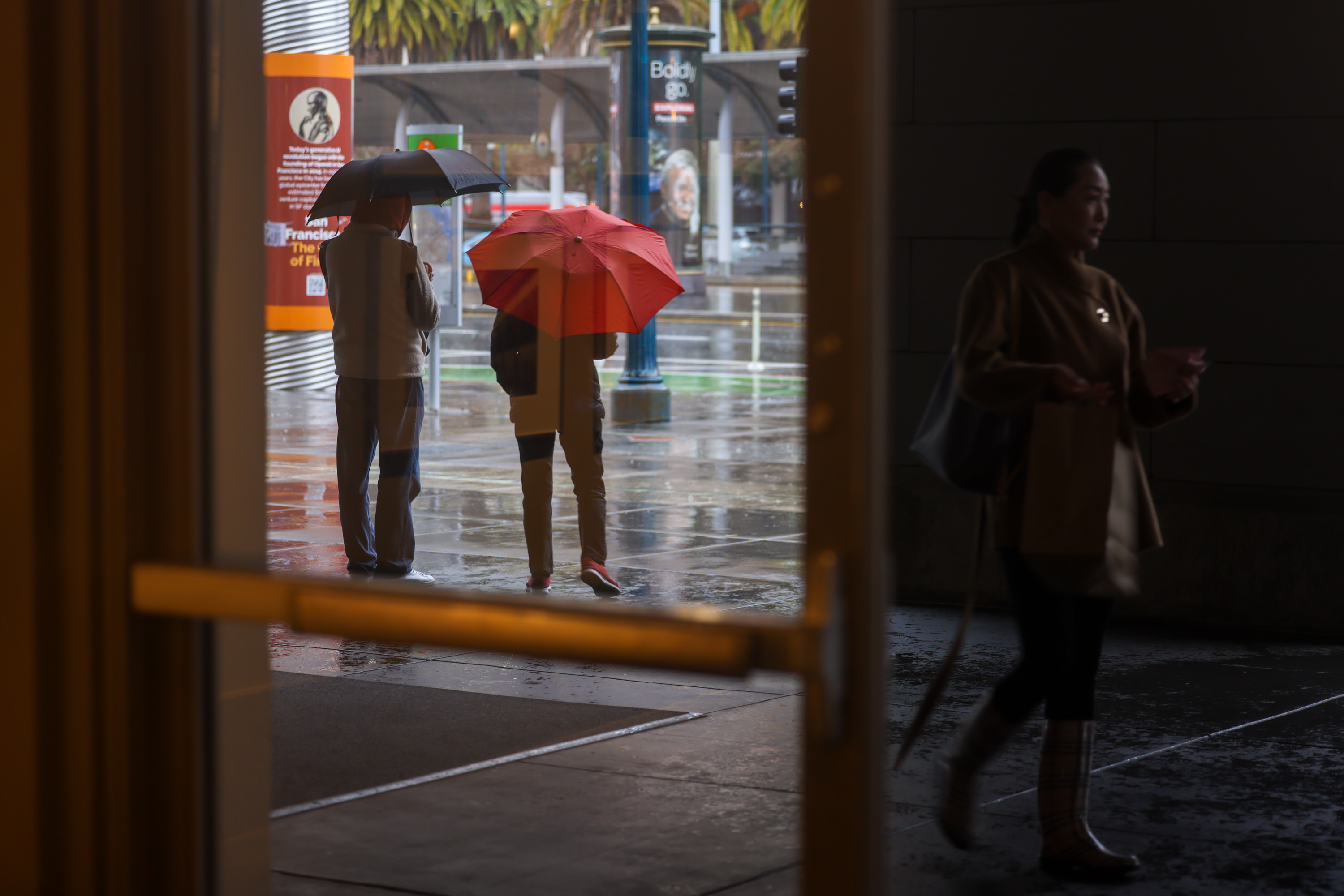 Two people stand on a sidewalk and hold umbrellas as they watch rain fall on a wet weather day.