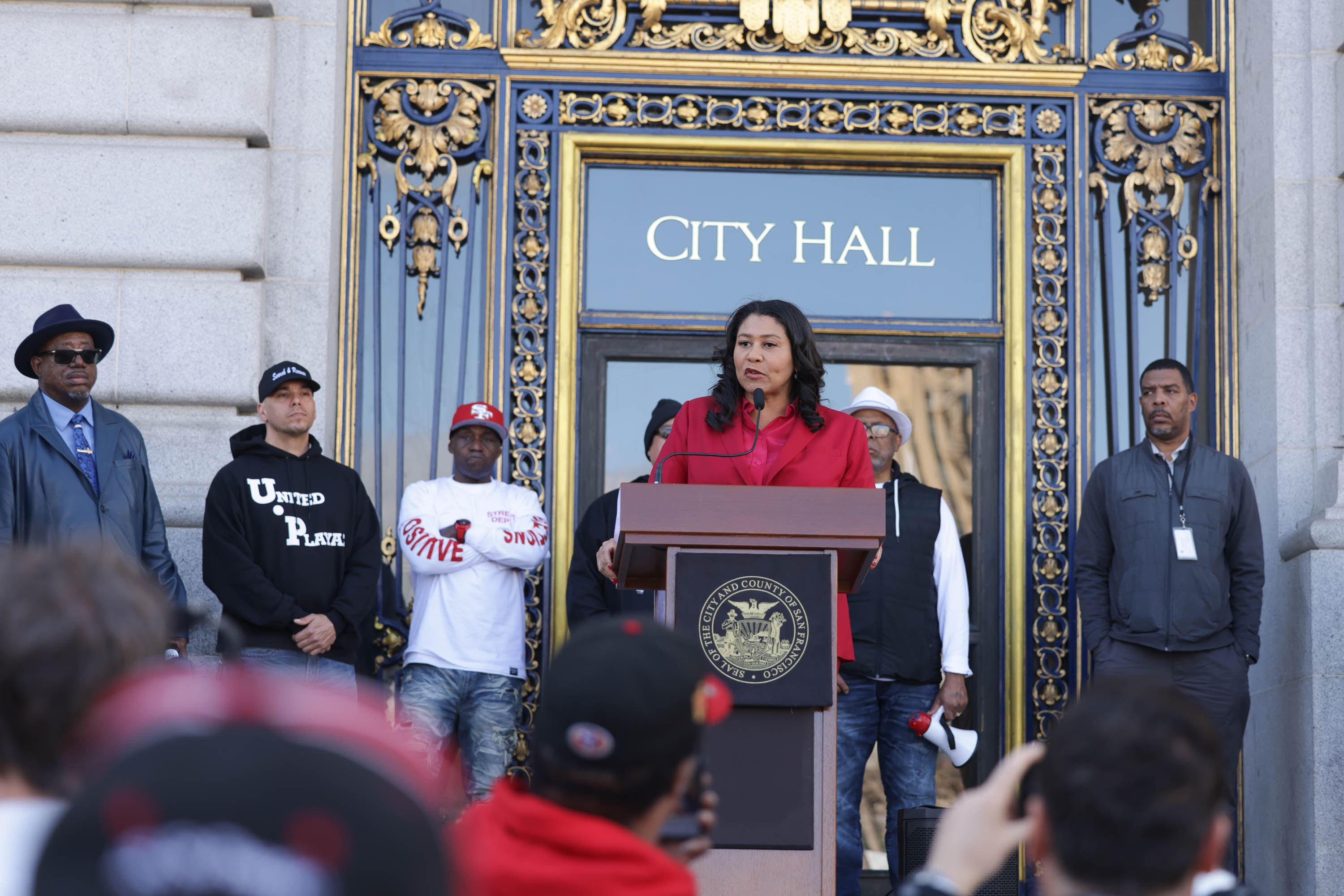 A woman speaks at a podium marked &quot;CITY HALL&quot; with onlookers behind her, some wearing shirts with &quot;UNITE&quot; and &quot;SAVE&quot; visible.