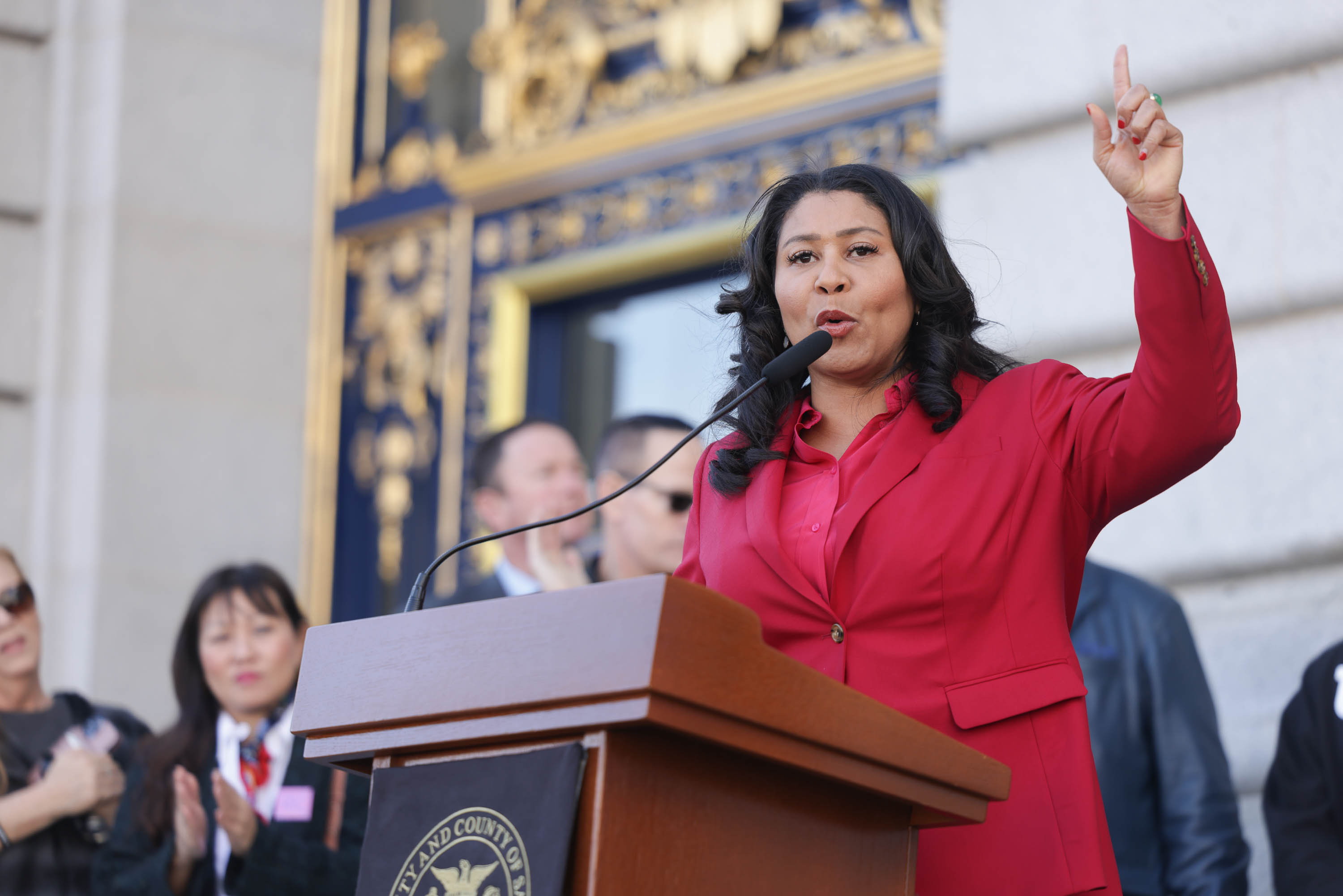 A woman in a red suit gestures energetically at a podium, addressing an audience, with others clapping in the background.