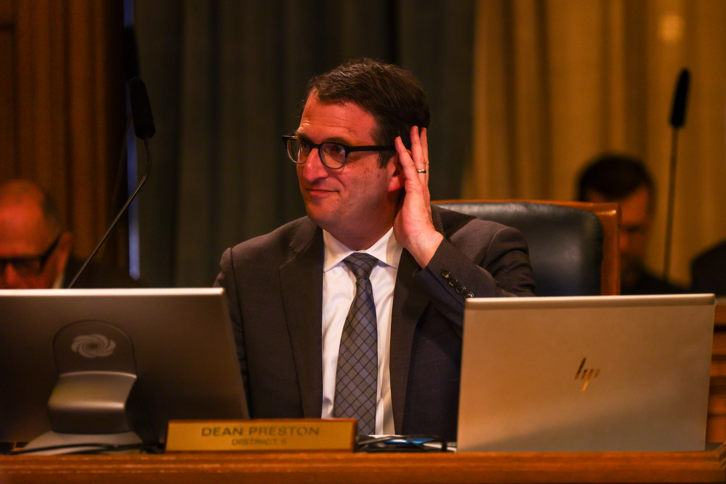 A man in a suit sits at a desk with two laptops, adjusts his glasses while looking to the side. A nameplate in front reads "Dean Preston, District 5."