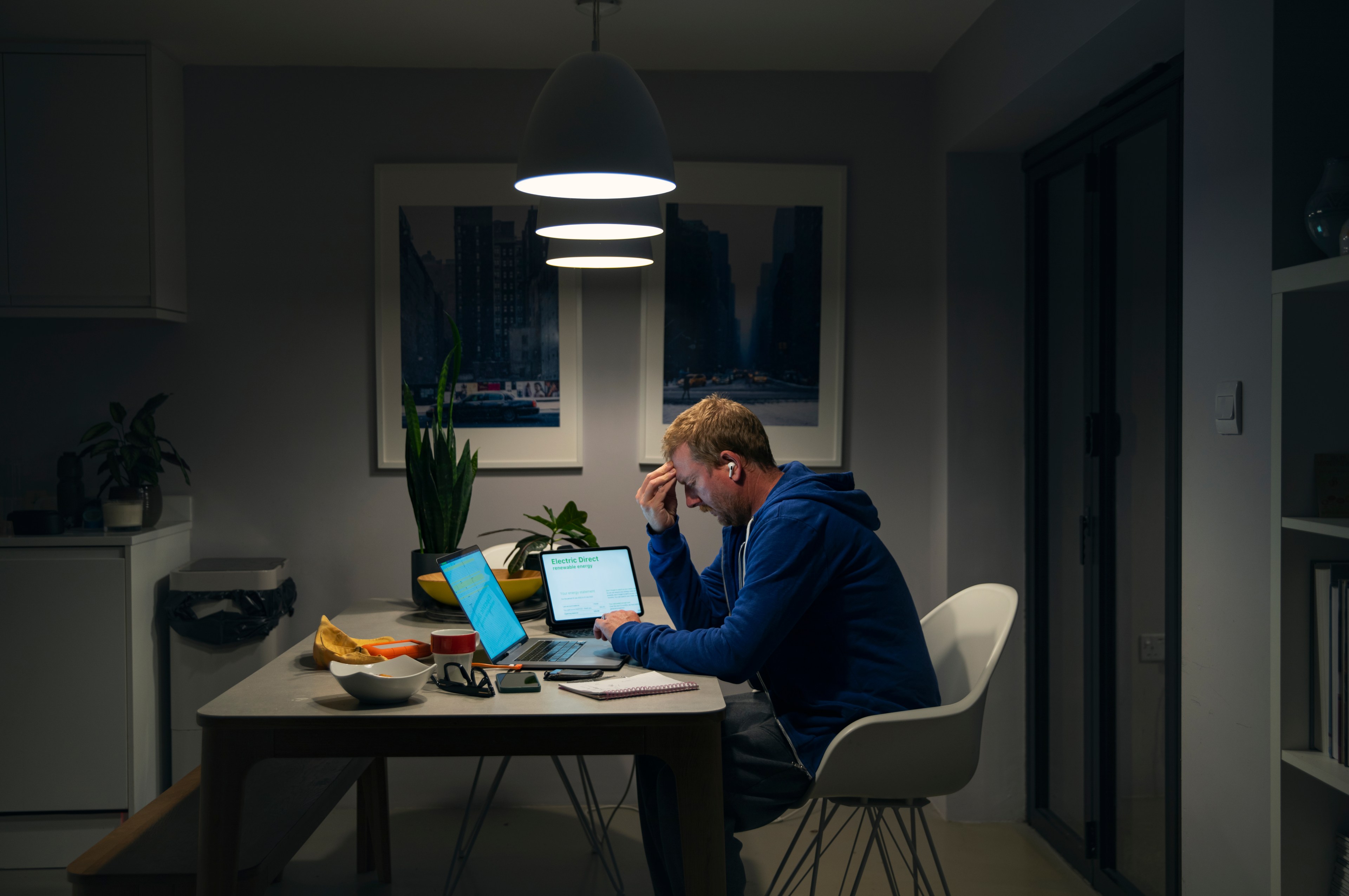 A person stressed at a home workspace at night, with a laptop, papers, and a lit lamp overhead.