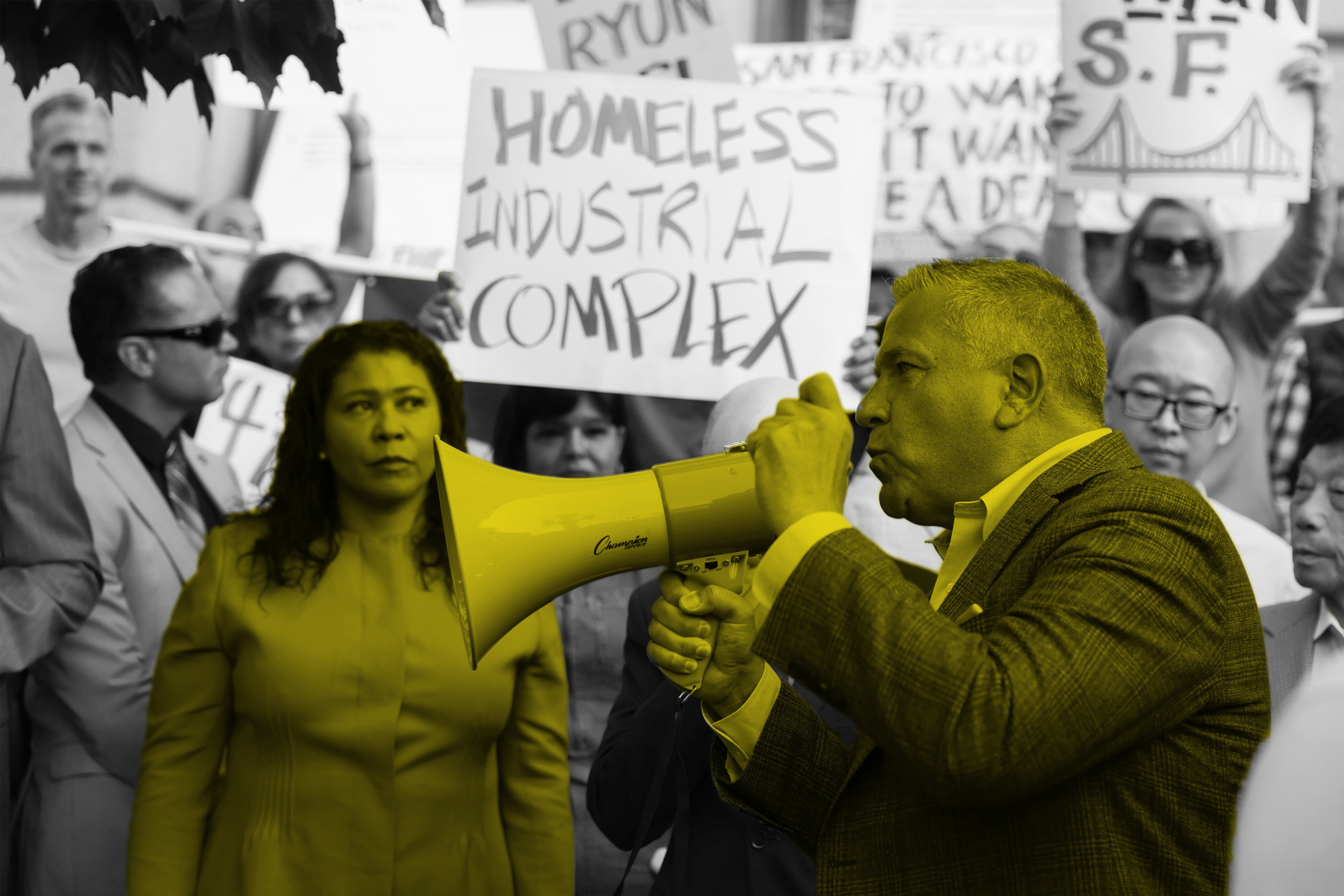 A man in a suit speaks into a yellow megaphone at a protest; black-and-white backdrop with only the megaphone in color.
