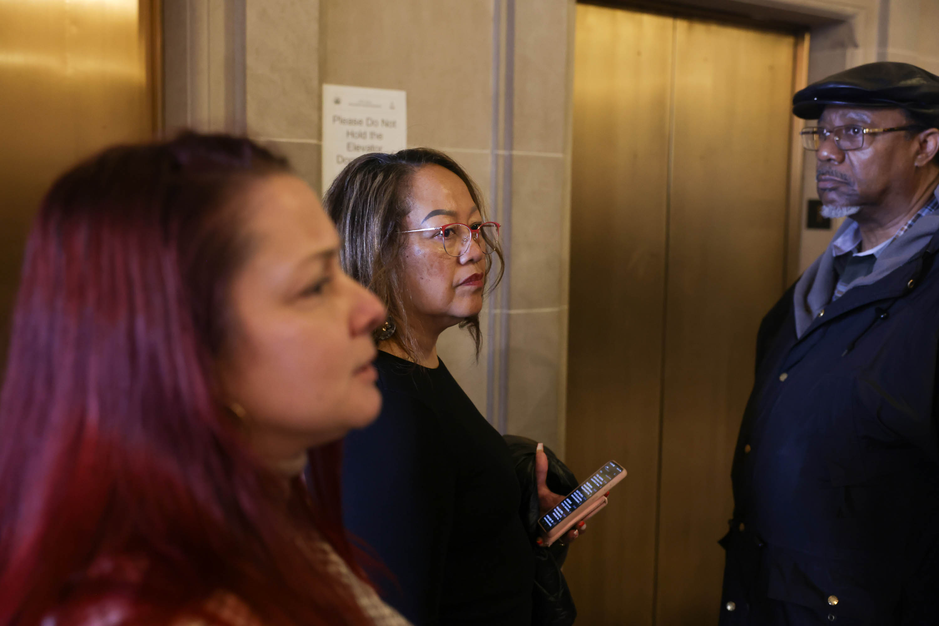 Three adults stand near an elevator, one holding a phone, with concerned expressions.