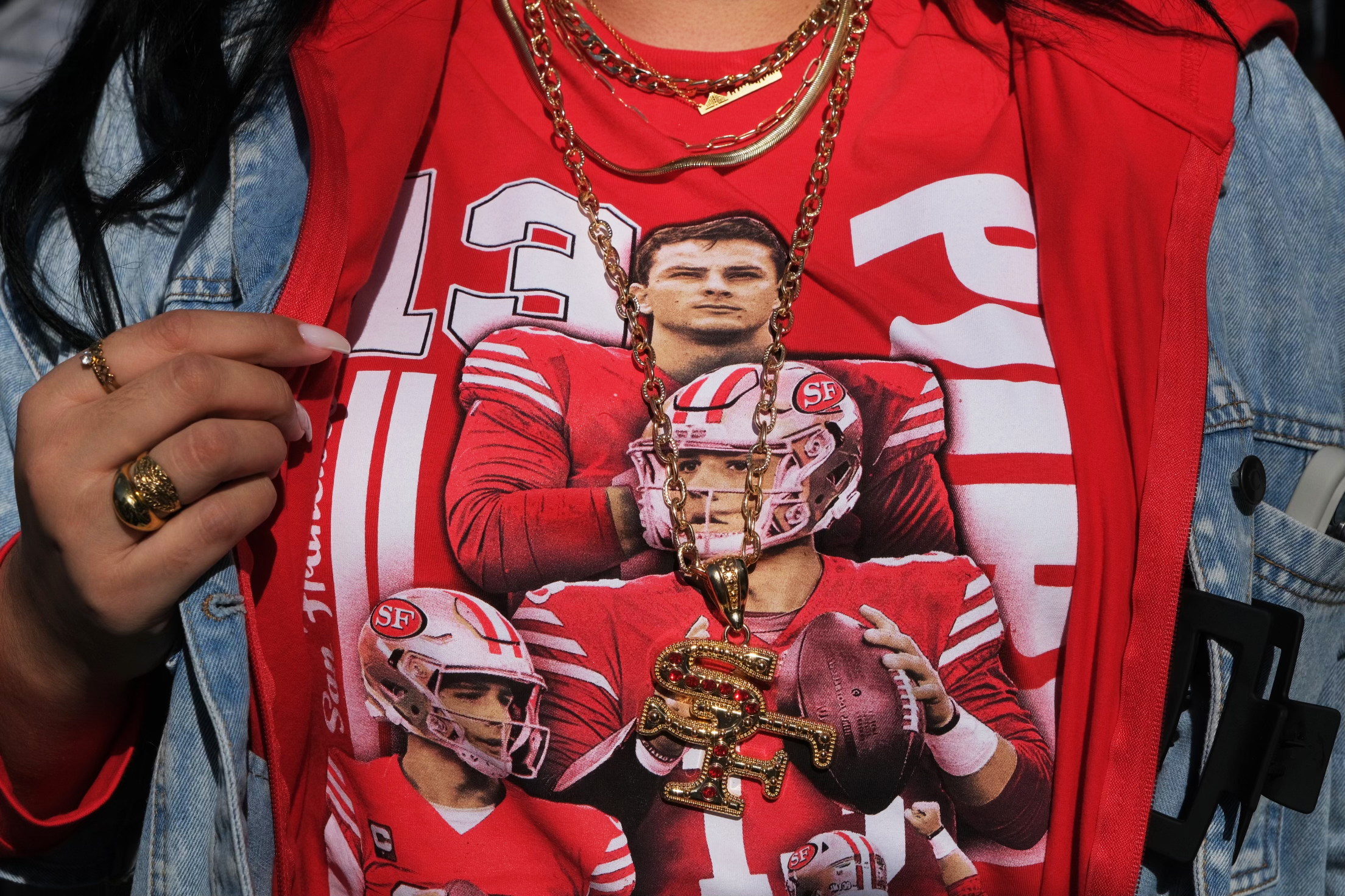 A person is wearing a red T-shirt with printed images of football players and the number 13, layered with a denim jacket and gold jewelry.