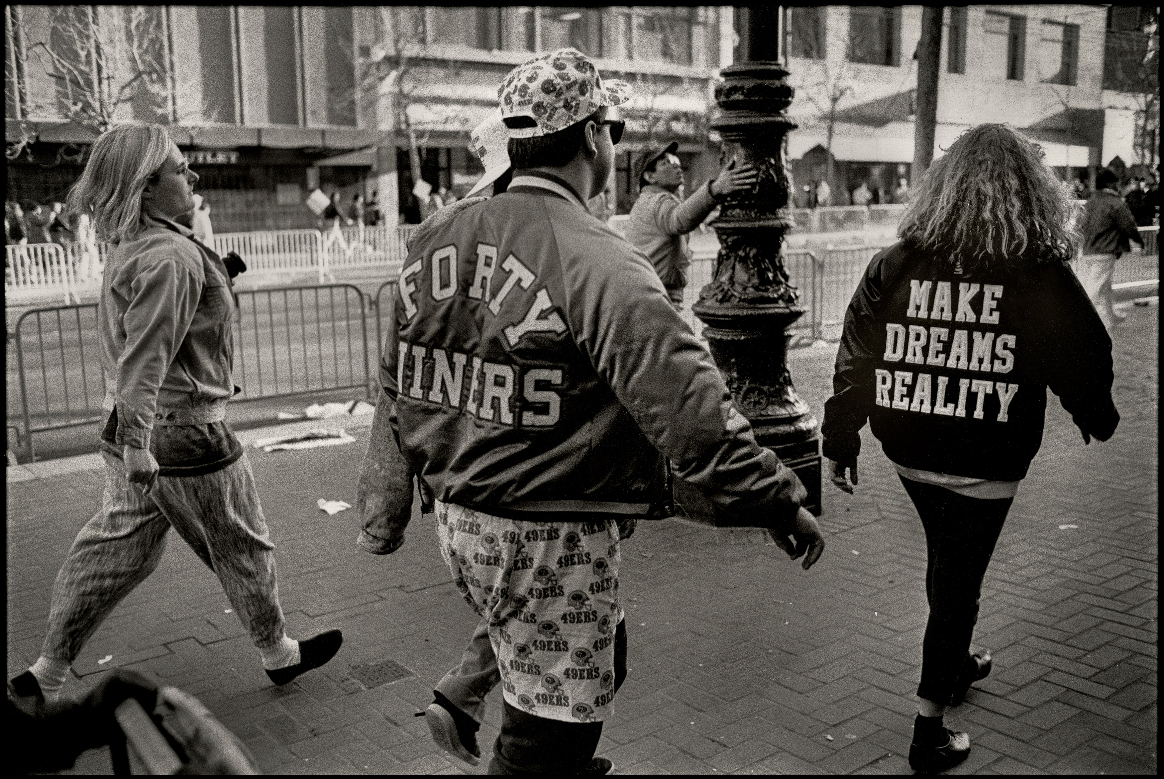 A black and white photo of a group of people, one wearing a vintage Forty-Niners jacket, walk along a street.