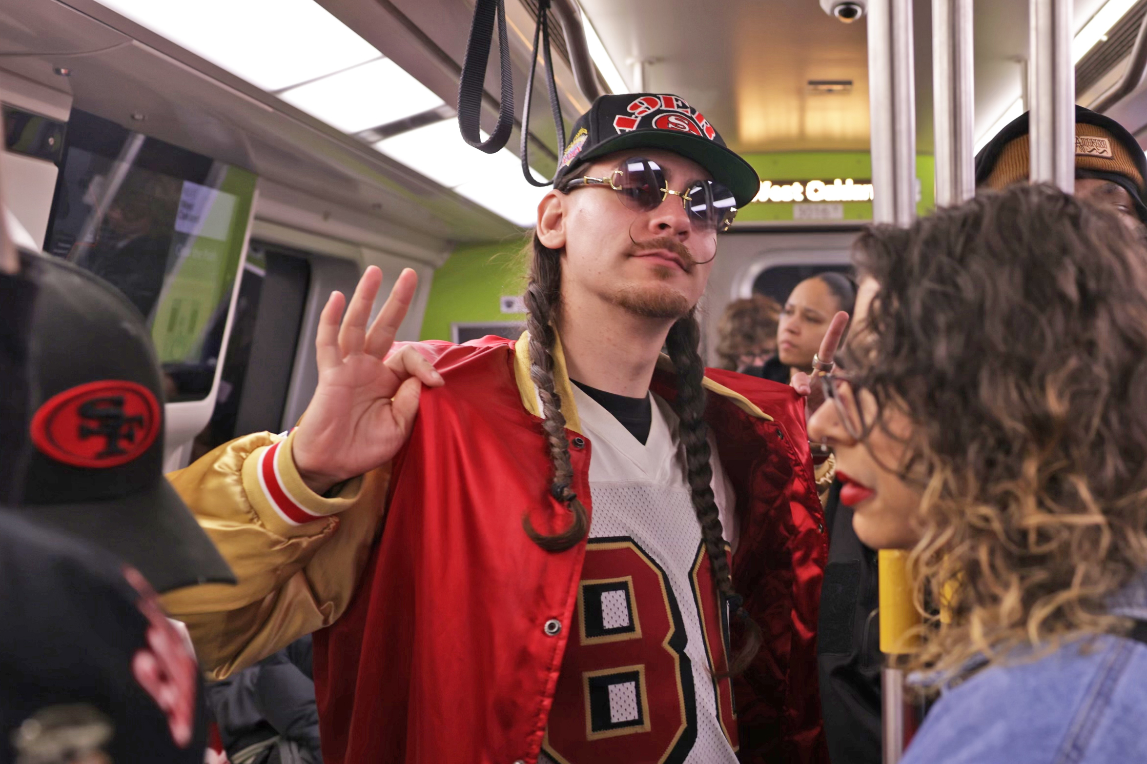 A man in a red and gold varsity jacket, wearing sunglasses, raises a peace sign on a crowded train.