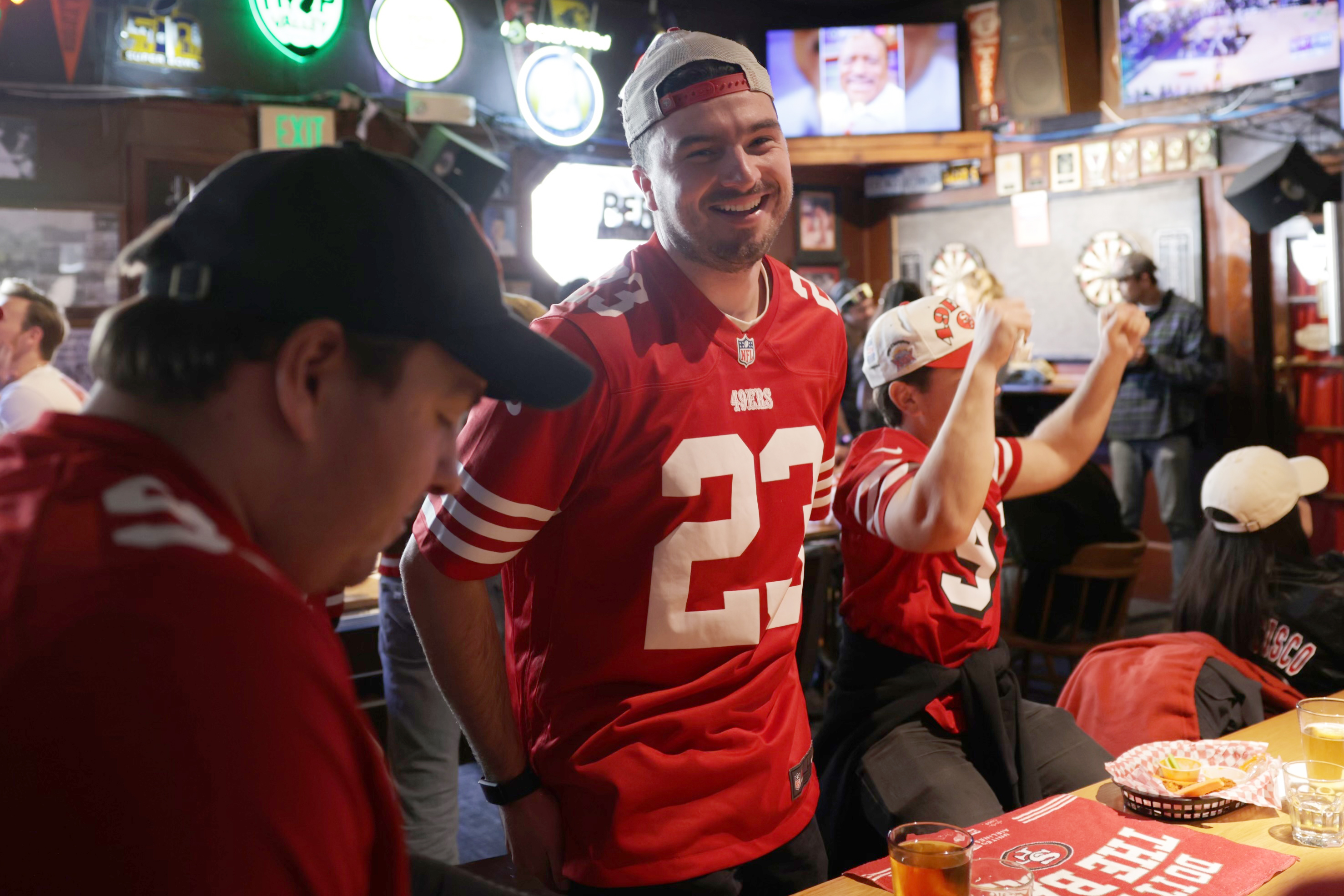 Man in 49ers jersey smiles inside bar as another man cheers behind him