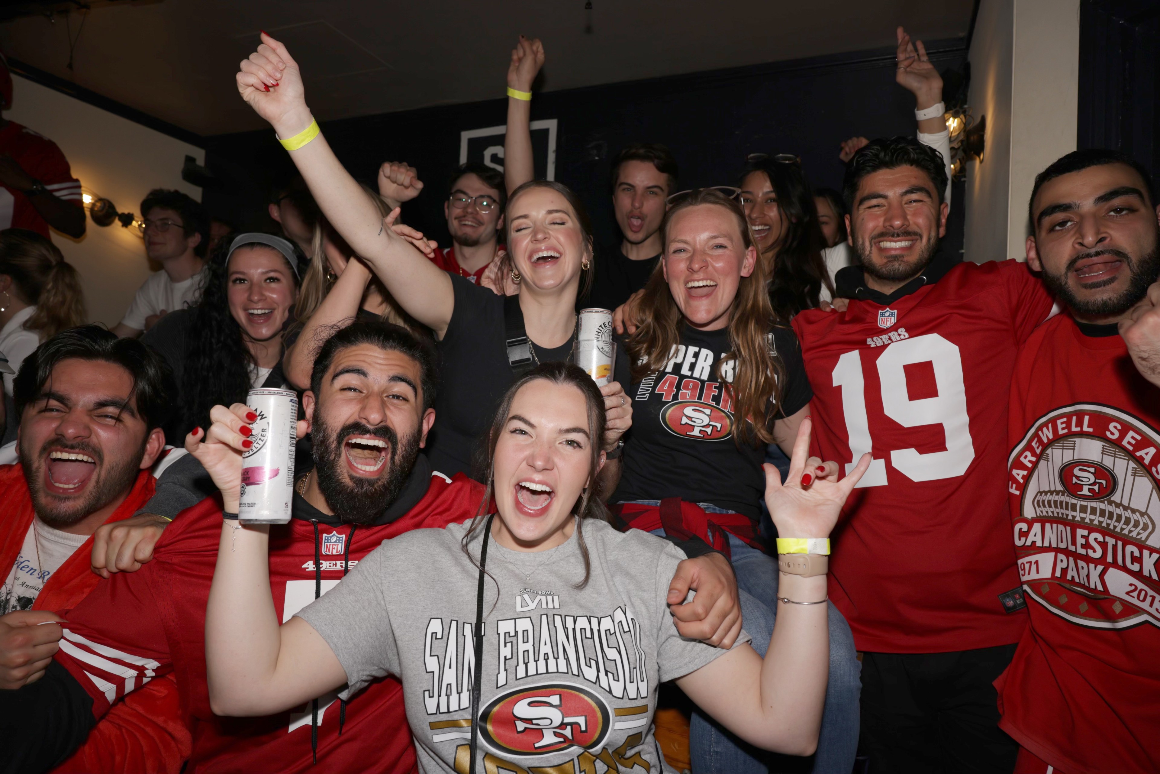 A group of jovial people in San Francisco 49ers gear, cheering with raised arms and holding drinks.