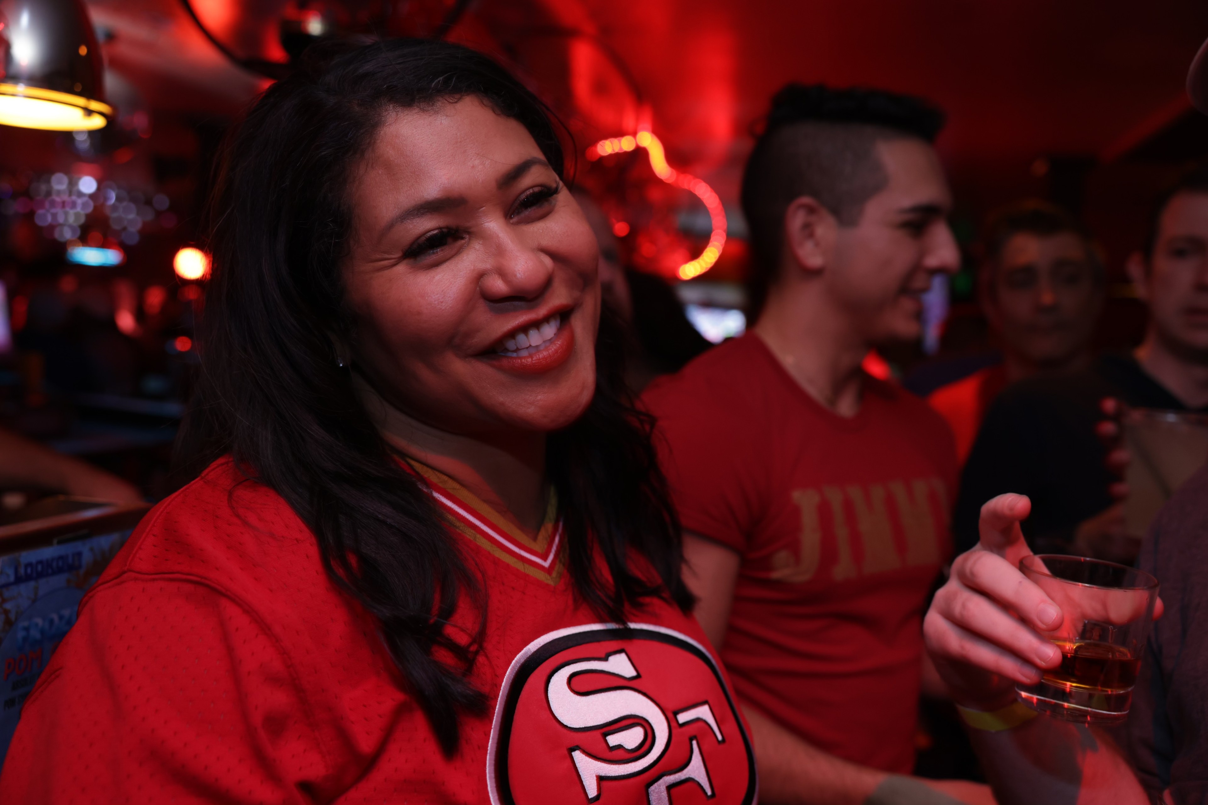 A smiling woman in a red sports jersey holds a drink in a bar with others nearby.