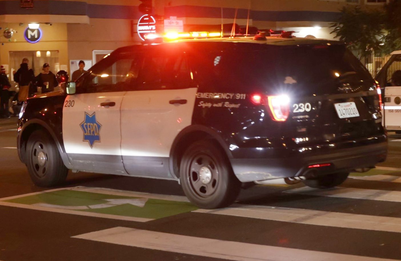 A police SUV with a lit siren is parked on a city street corner at night. The vehicle has "SFPD" on the side, and people are gathered in the background.