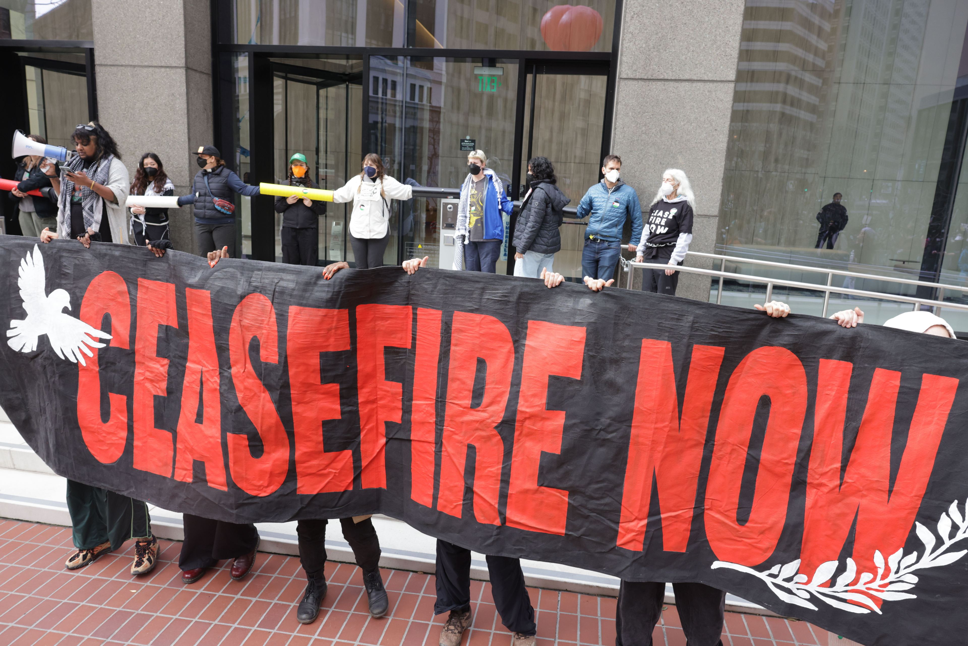 Protesters hold a black banner with red lettering outside a downtown building.