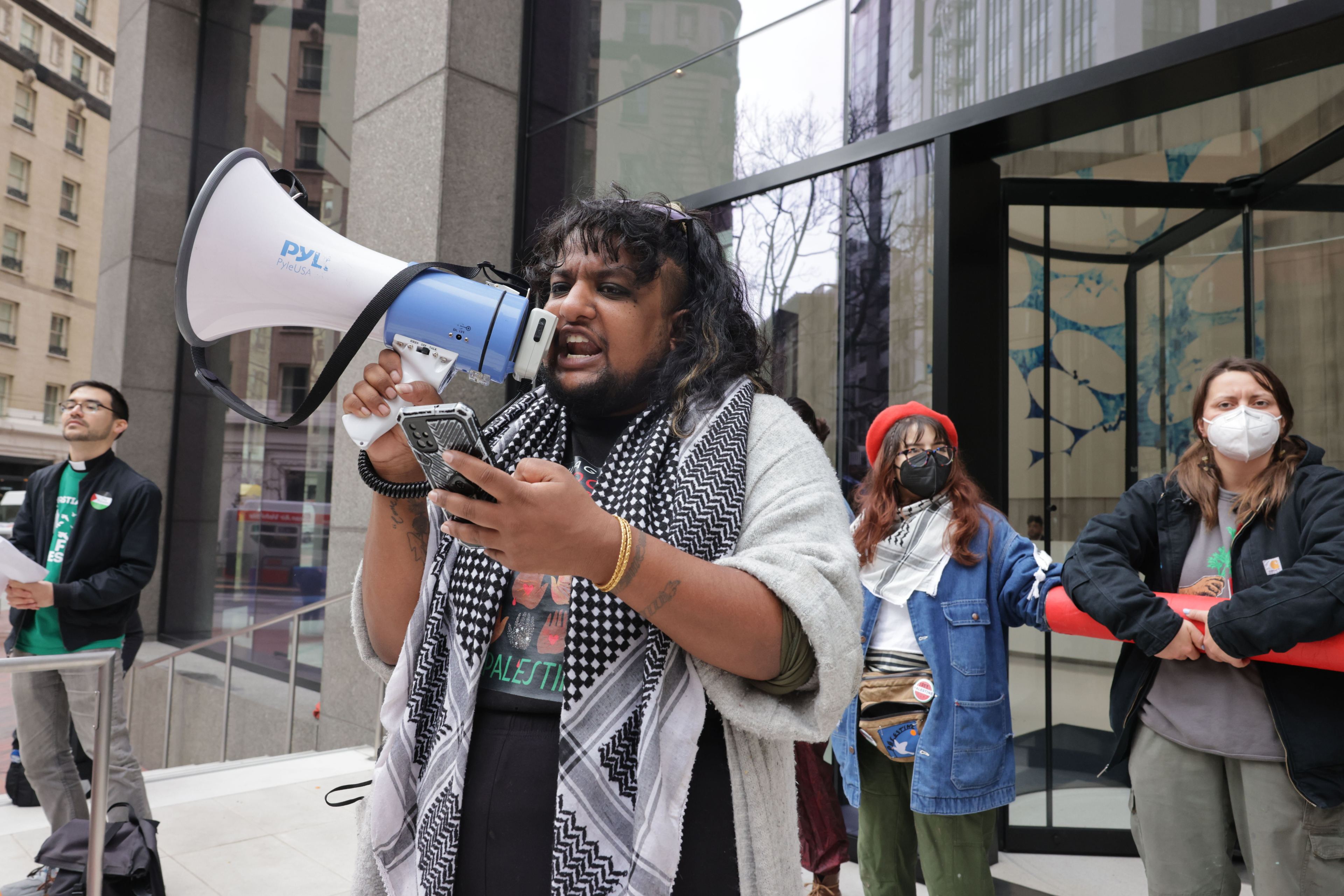 A person holding a bullhorn reads a statement from a cellphone in their hand outside an office building.